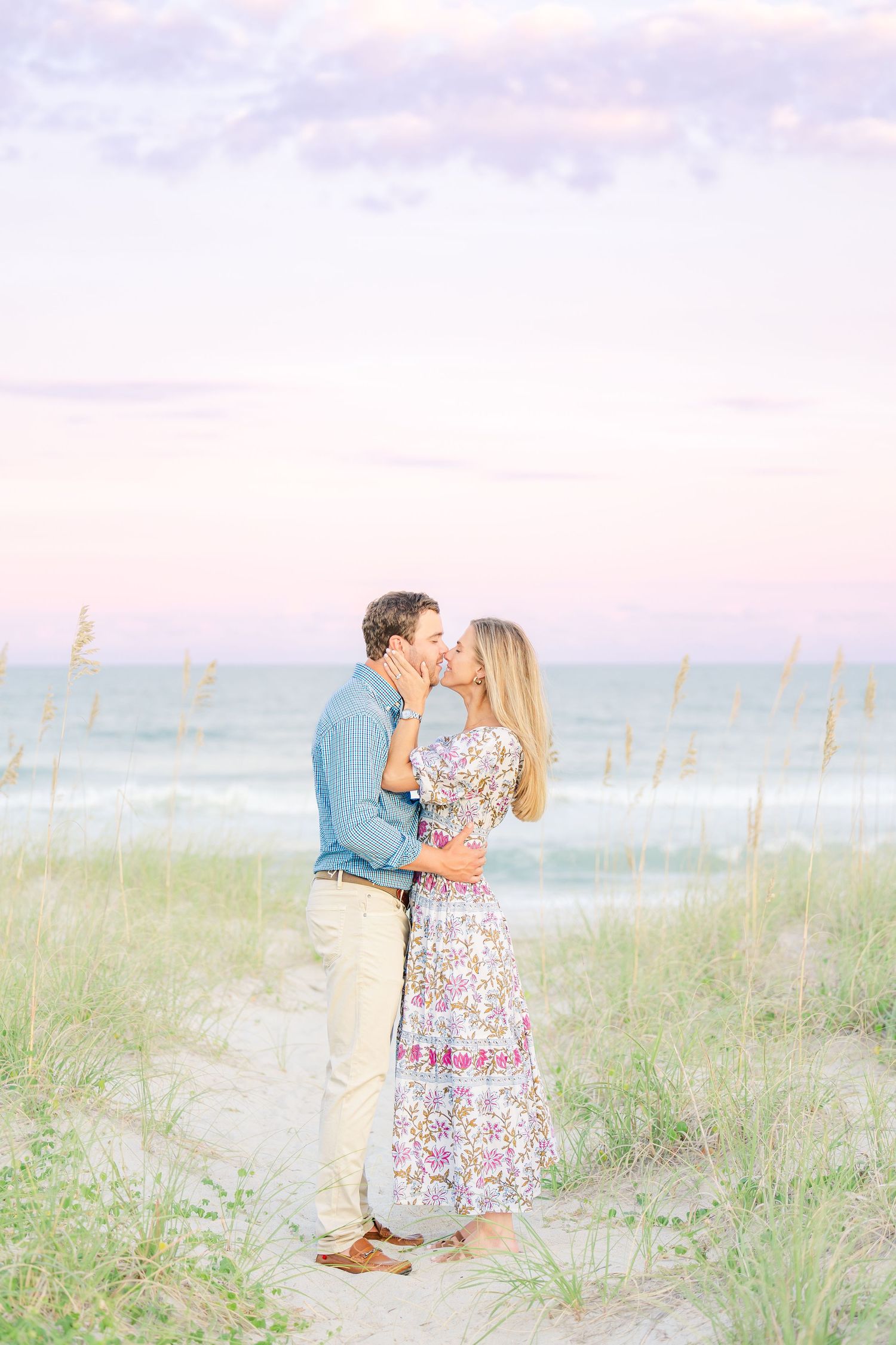 Loving embrace on coastal path with ocean waves and dune grass in soft lighting.