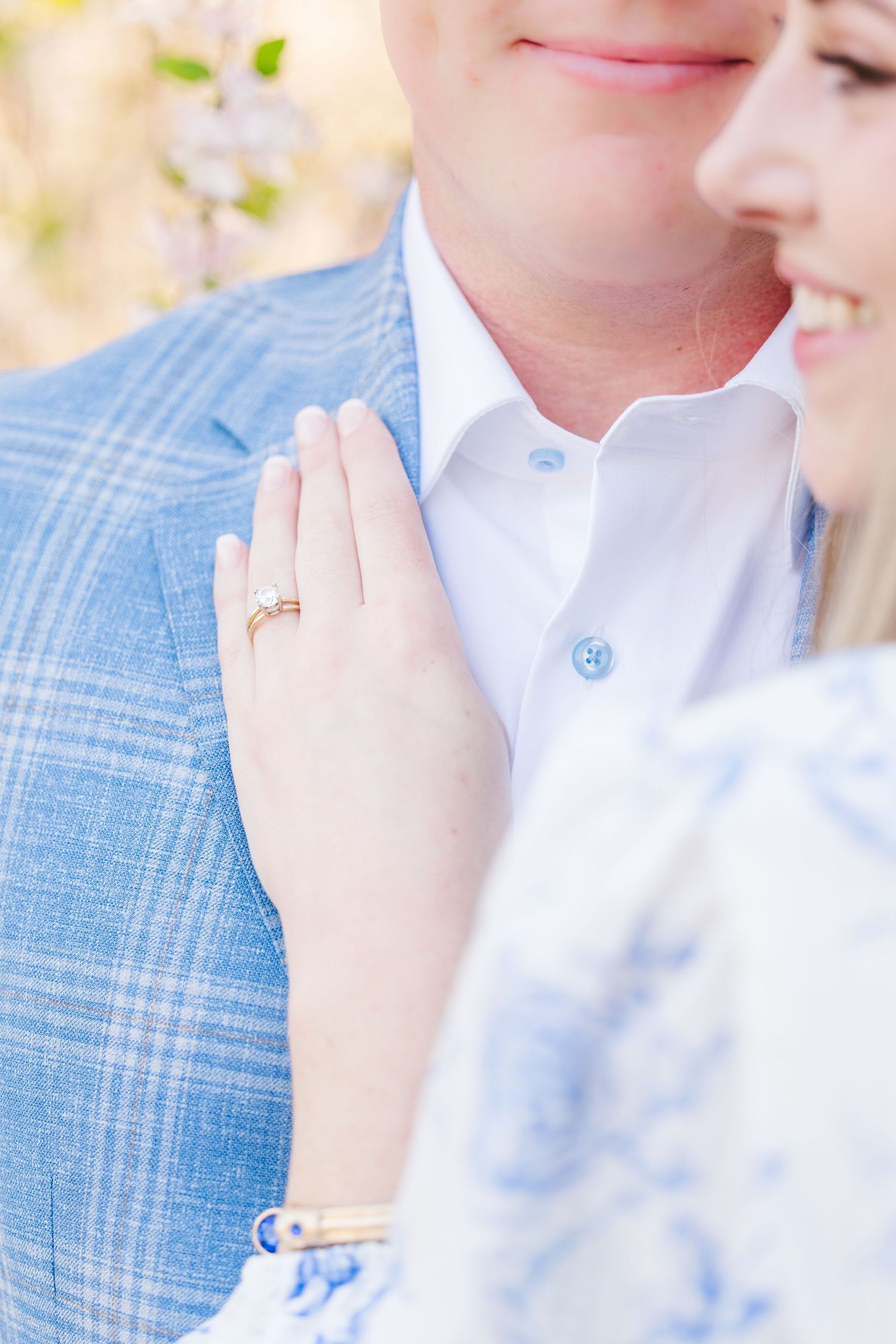 Close up of a couple showing an engagement ring with a light blue plaid suit jacket and white dress shirt.