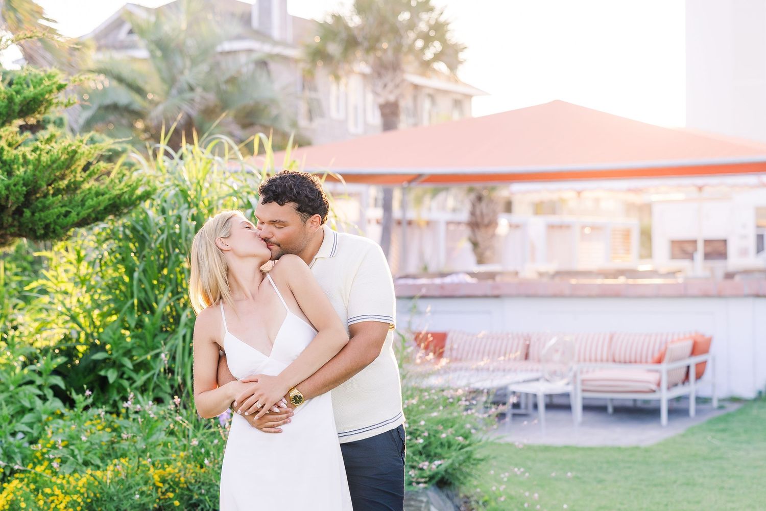 Couple sharing a romantic kiss in a lush garden setting with tropical plants and resort architecture in the background.