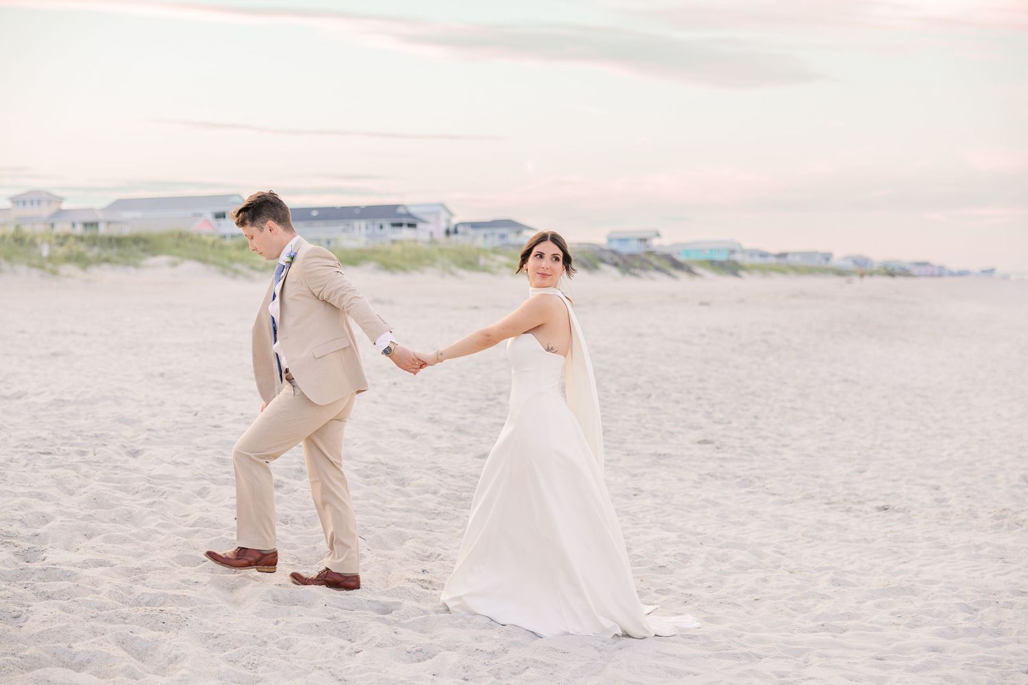 Bride and groom walking hand in hand on sandy beach at sunset in wedding attire in Carolina Beach near Wilmington, NC.