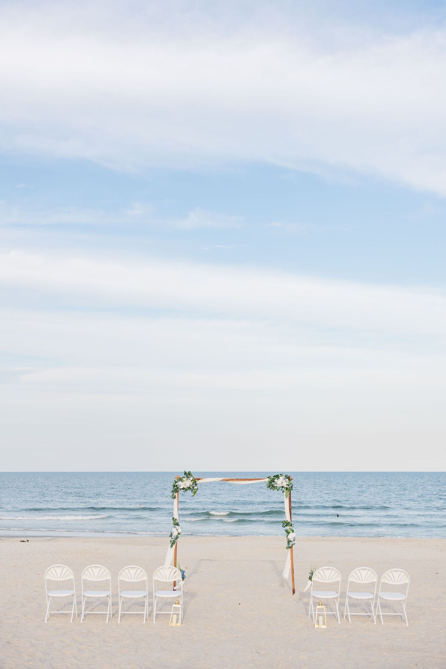 Elegant beach wedding setup with wooden floral arch, white chairs, and ocean backdrop under blue sky in Carolina Beach near Wilmington, NC.