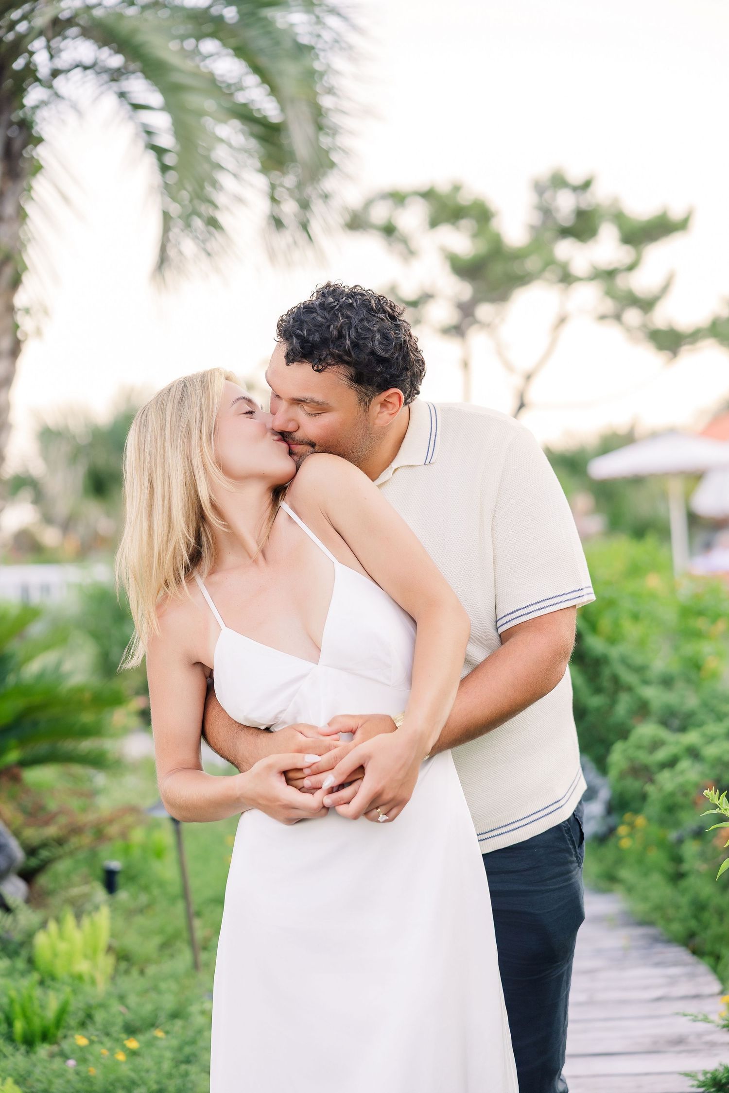 Couple sharing a tender kiss on a garden pathway surrounded by palm trees at dusk.