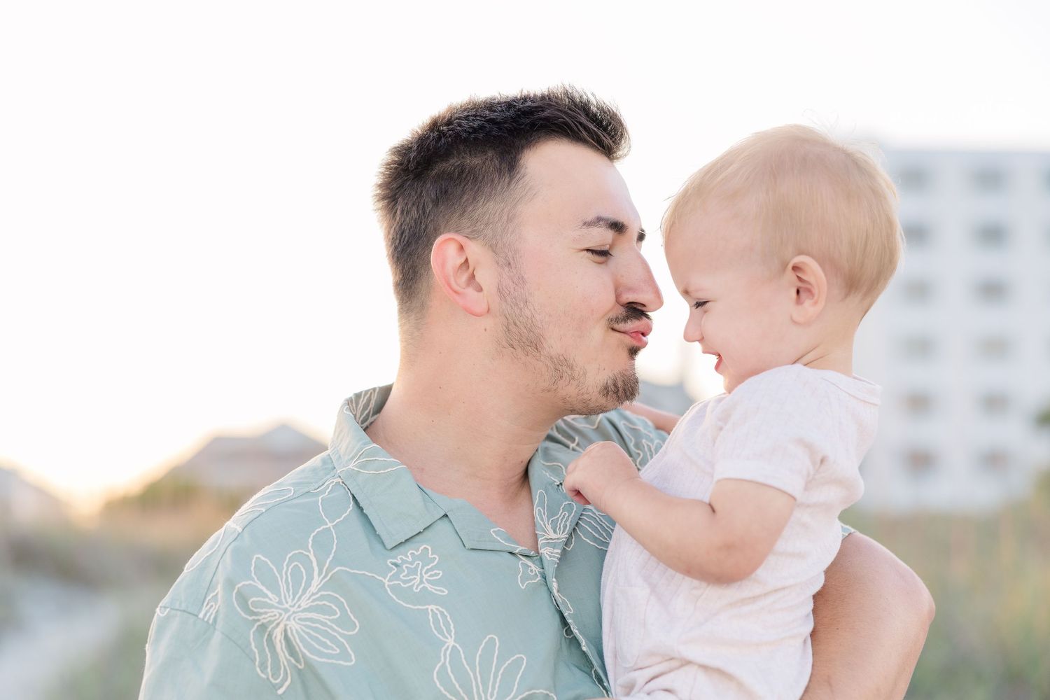 A loving father holds his baby close, touching noses and smiling on a bright sunny beach day.