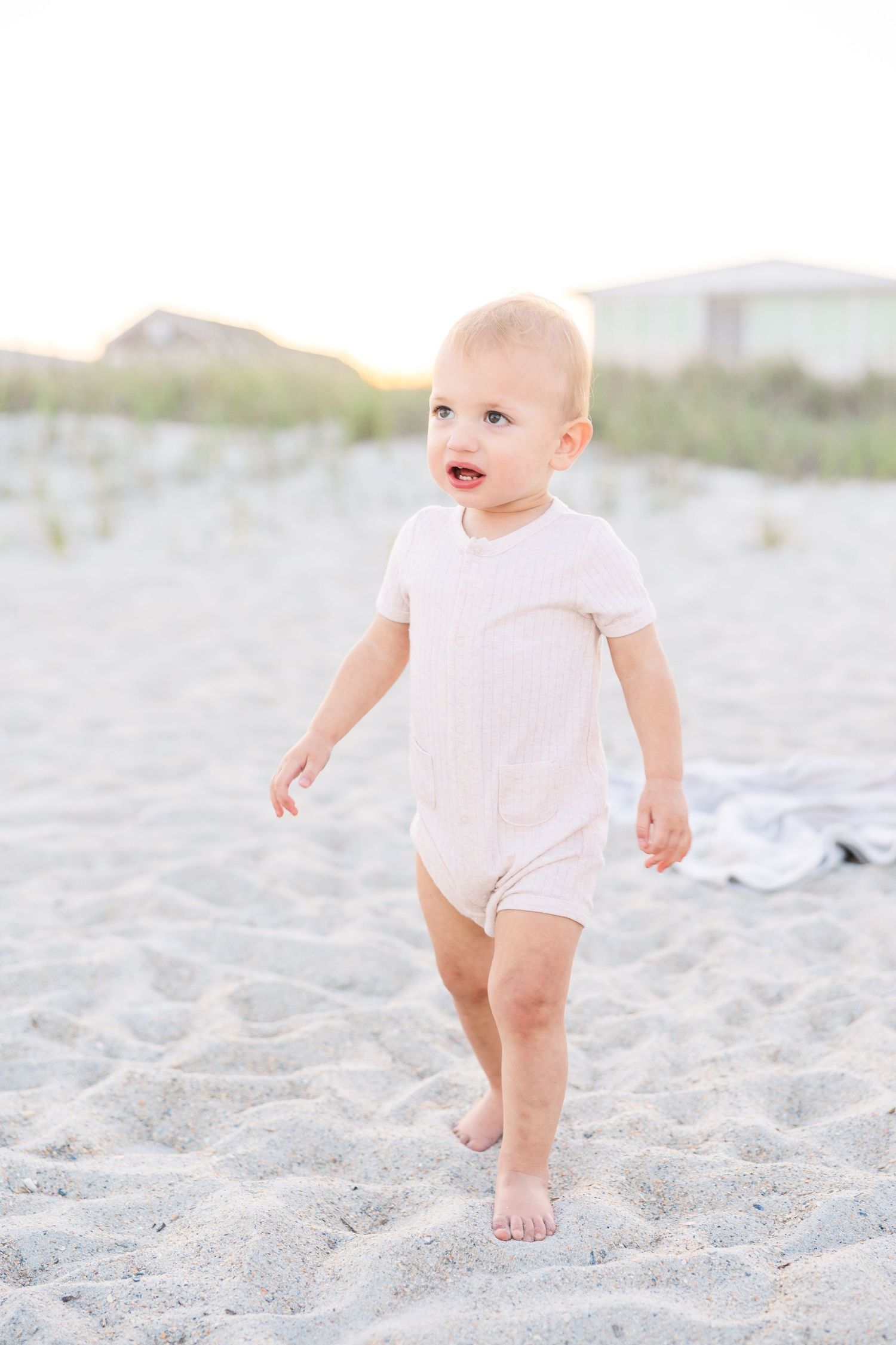 A happy toddler in a pink romper walks barefoot on sandy beach at sunset.