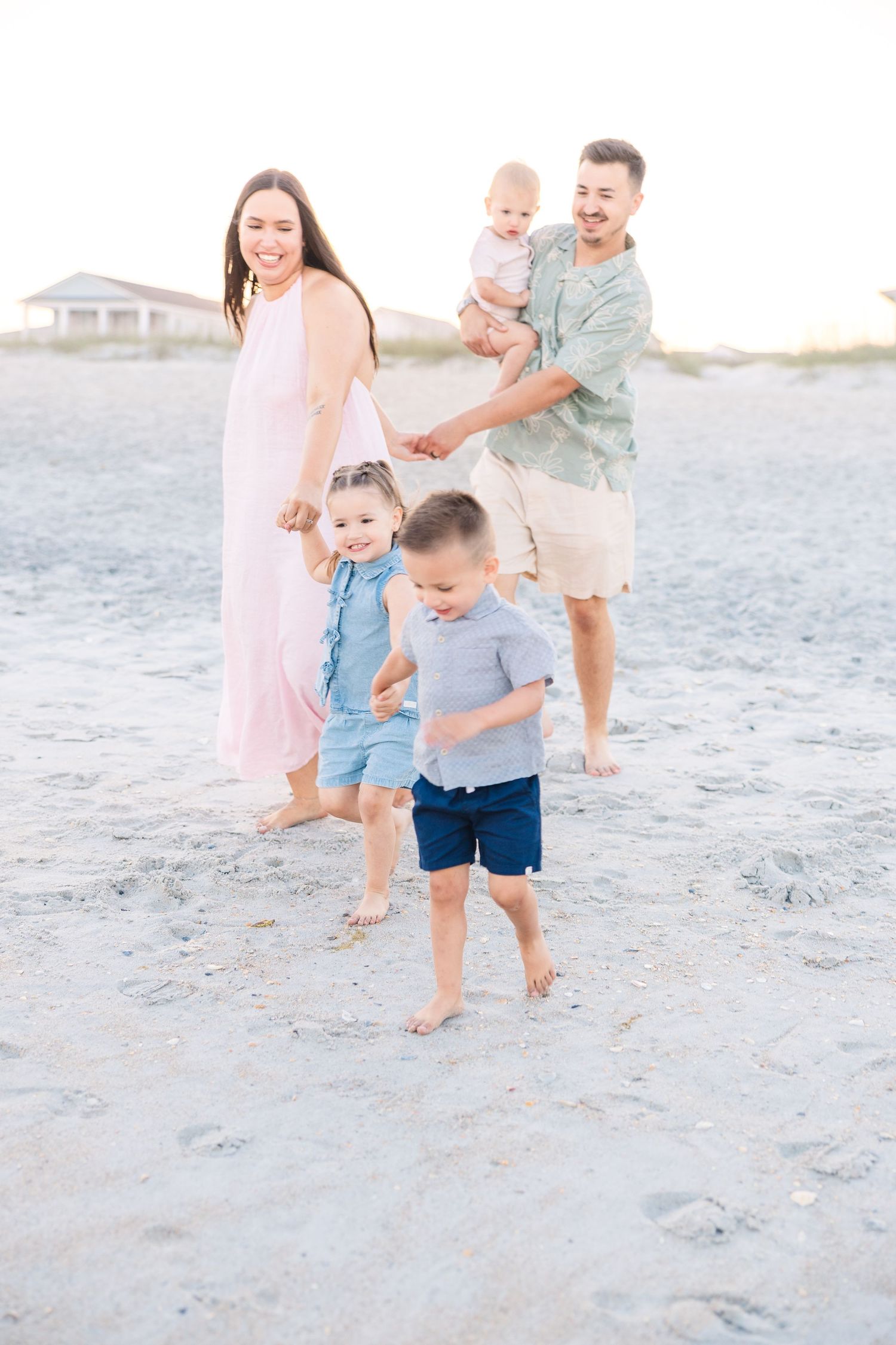 Family of five walking barefoot on sandy beach at sunset, parents holding children's hands.