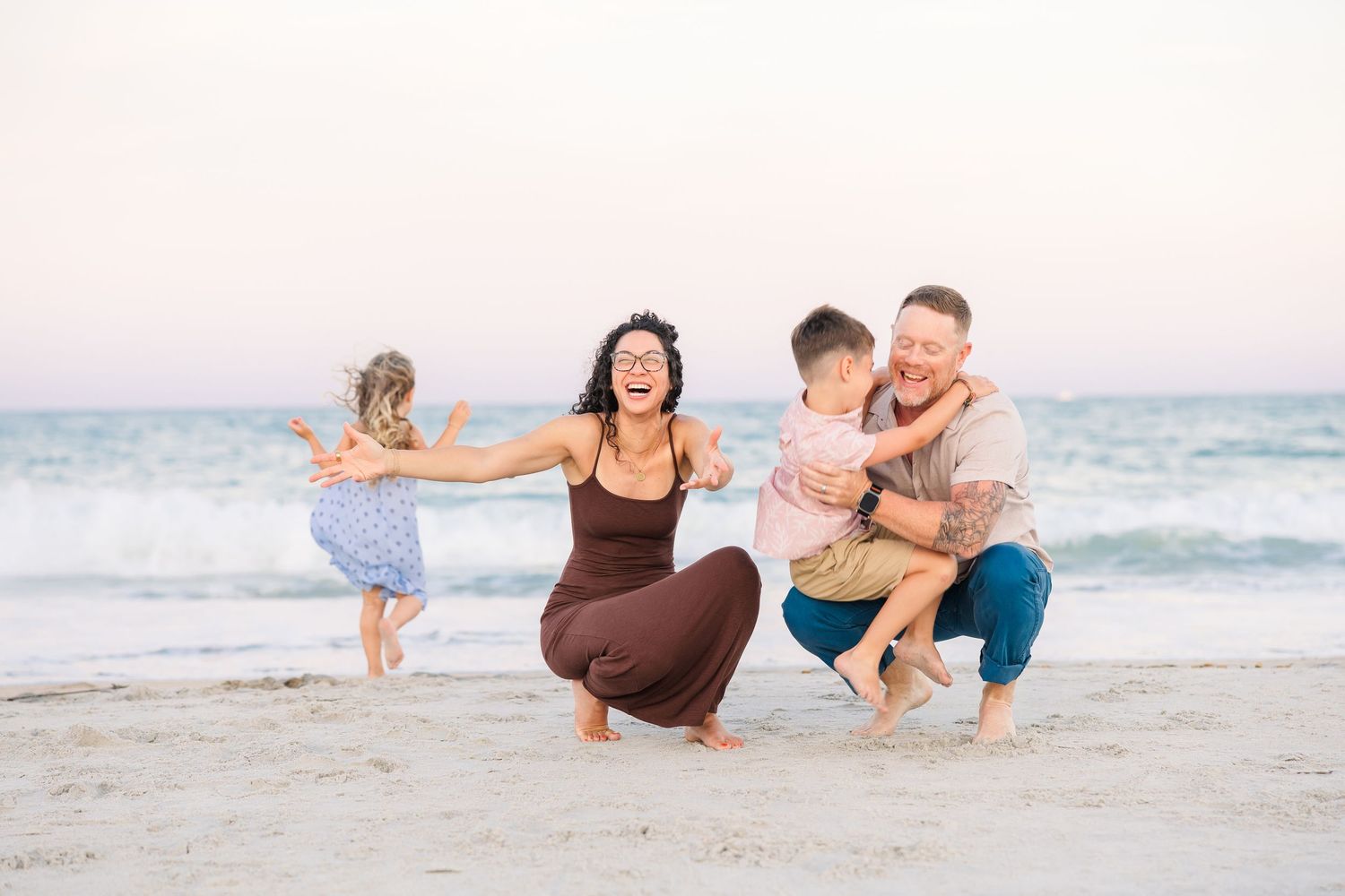 Happy family of four enjoying a sunny beach day, parents laughing with young children on sandy shore near ocean waves.