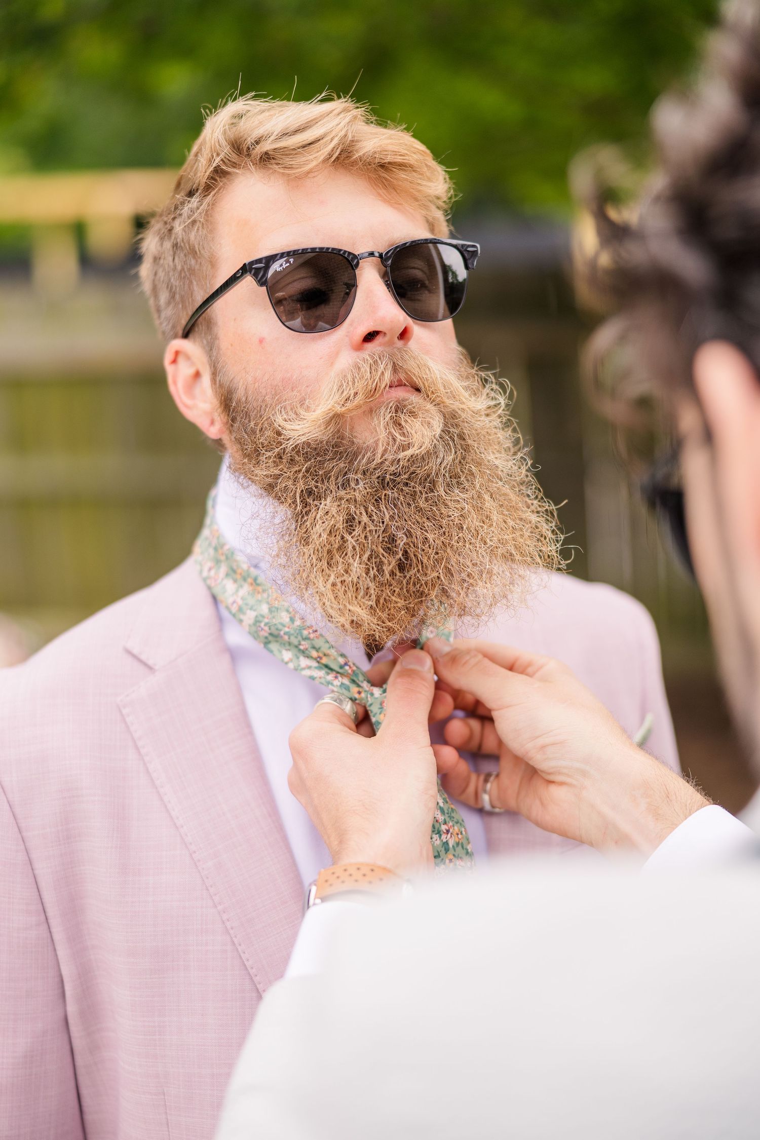 A groom with a full beard wearing sunglasses and pink suit jacket in an outdoor setting in a backyard wedding in wilmington, nc