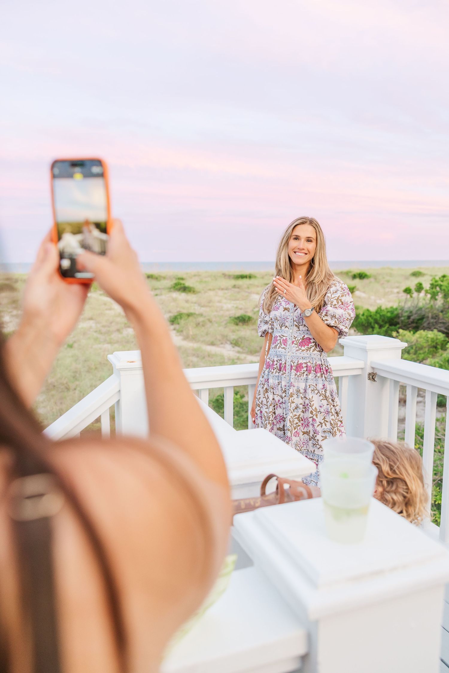 Someone takes a phone photo of a woman in a floral dress on a coastal deck.