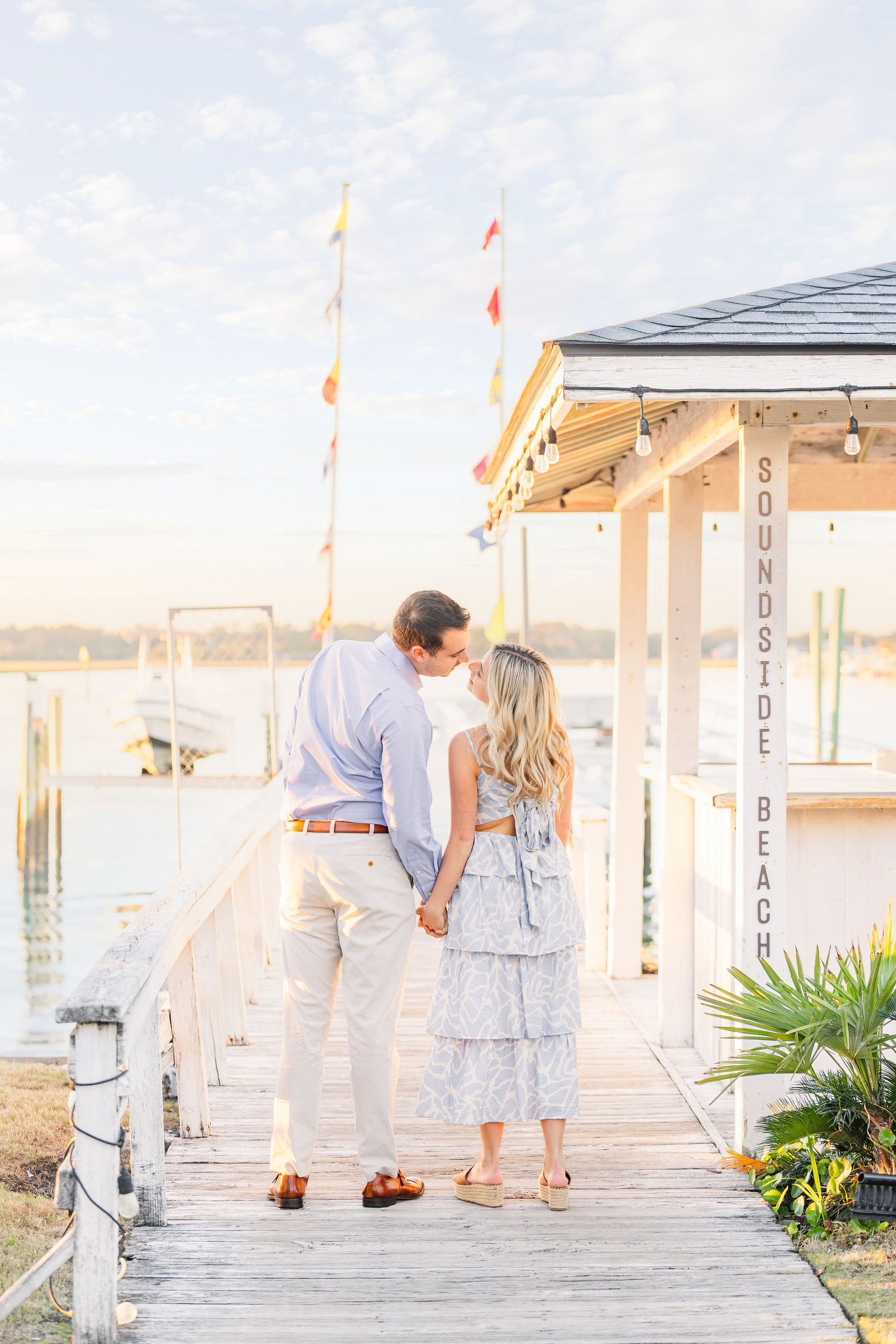 Romantic couple embraces on a coastal boardwalk at sunset near a beach house with nautical flags.