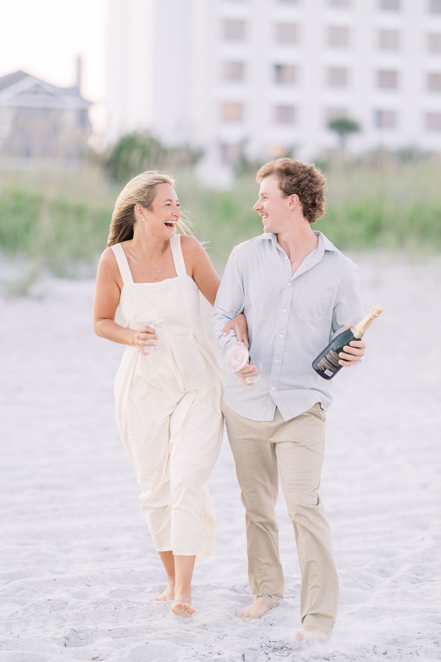 Couple walking and laughing together on beach with champagne bottle.