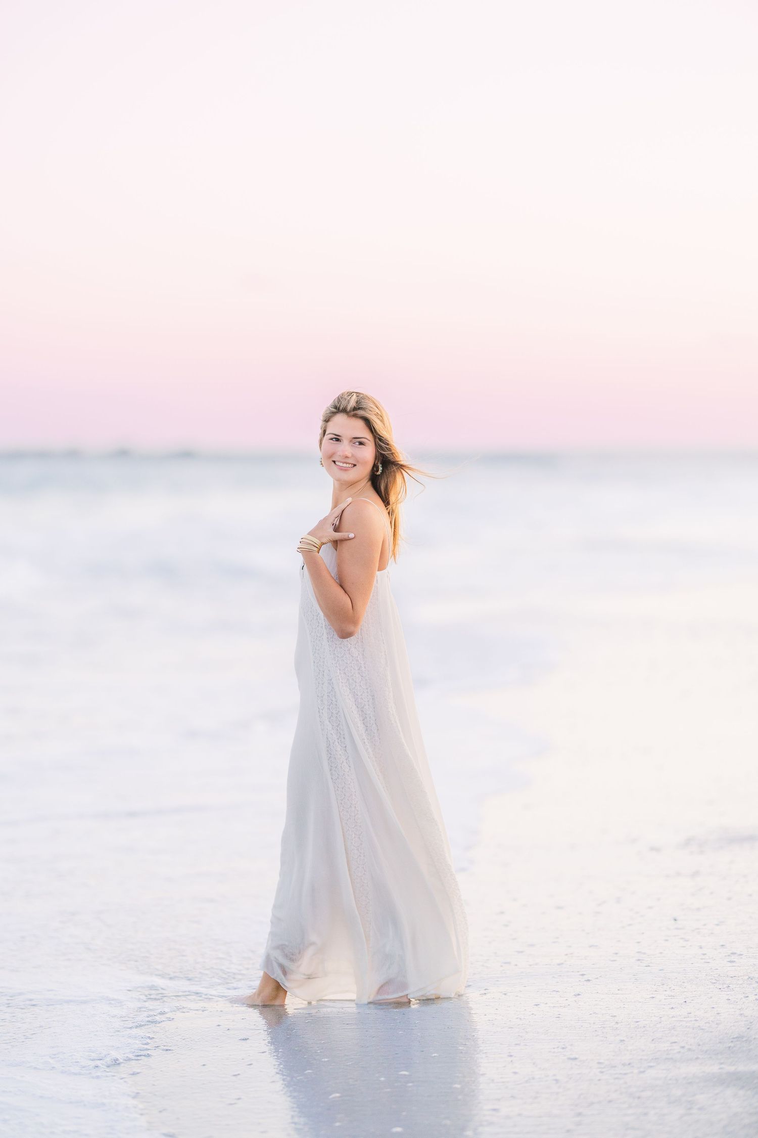 A dreamy beach photoshoot featuring a flowing white dress against soft pastel sunset colors.