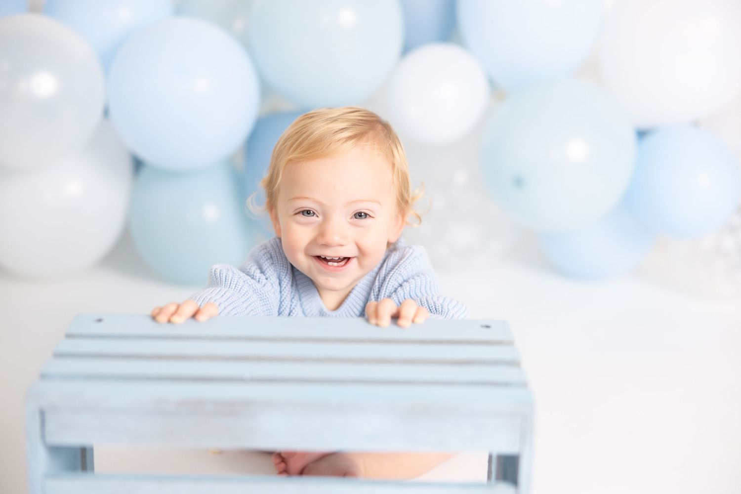 Joyful baby peeks over blue wooden crate with balloon decoration.