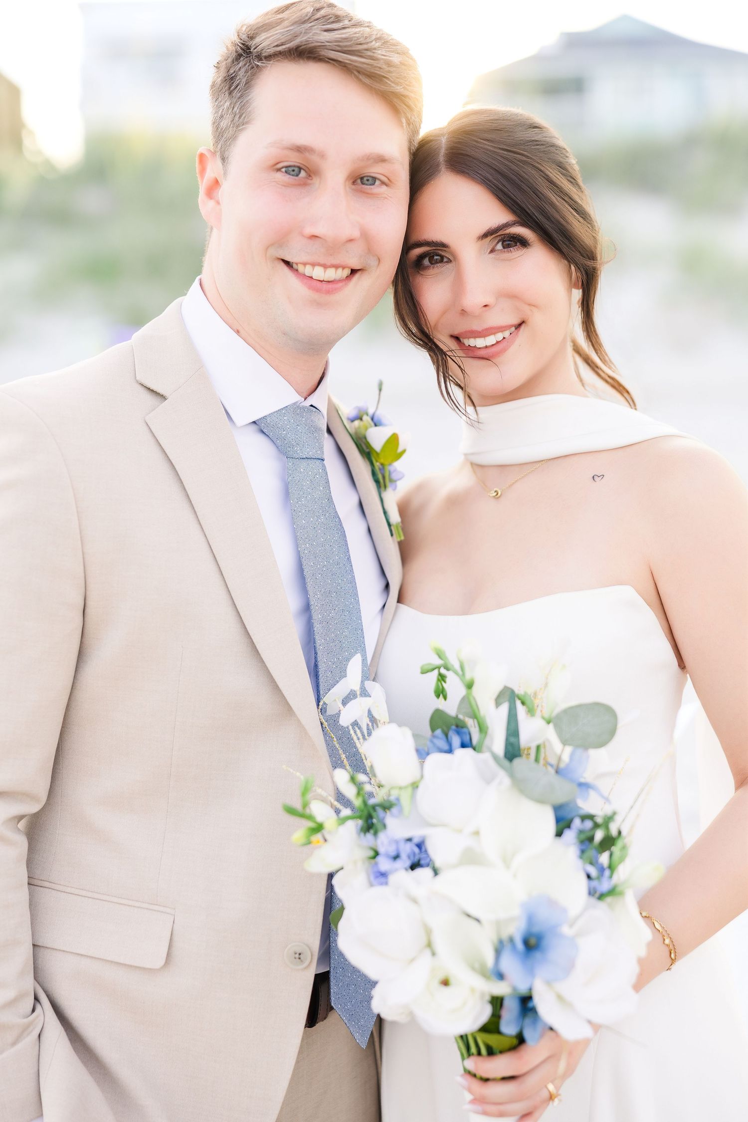 Smiling bride and groom in tan suit pose closely together holding white and blue floral bouquet on beach in Carolina Beach near Wilmington,