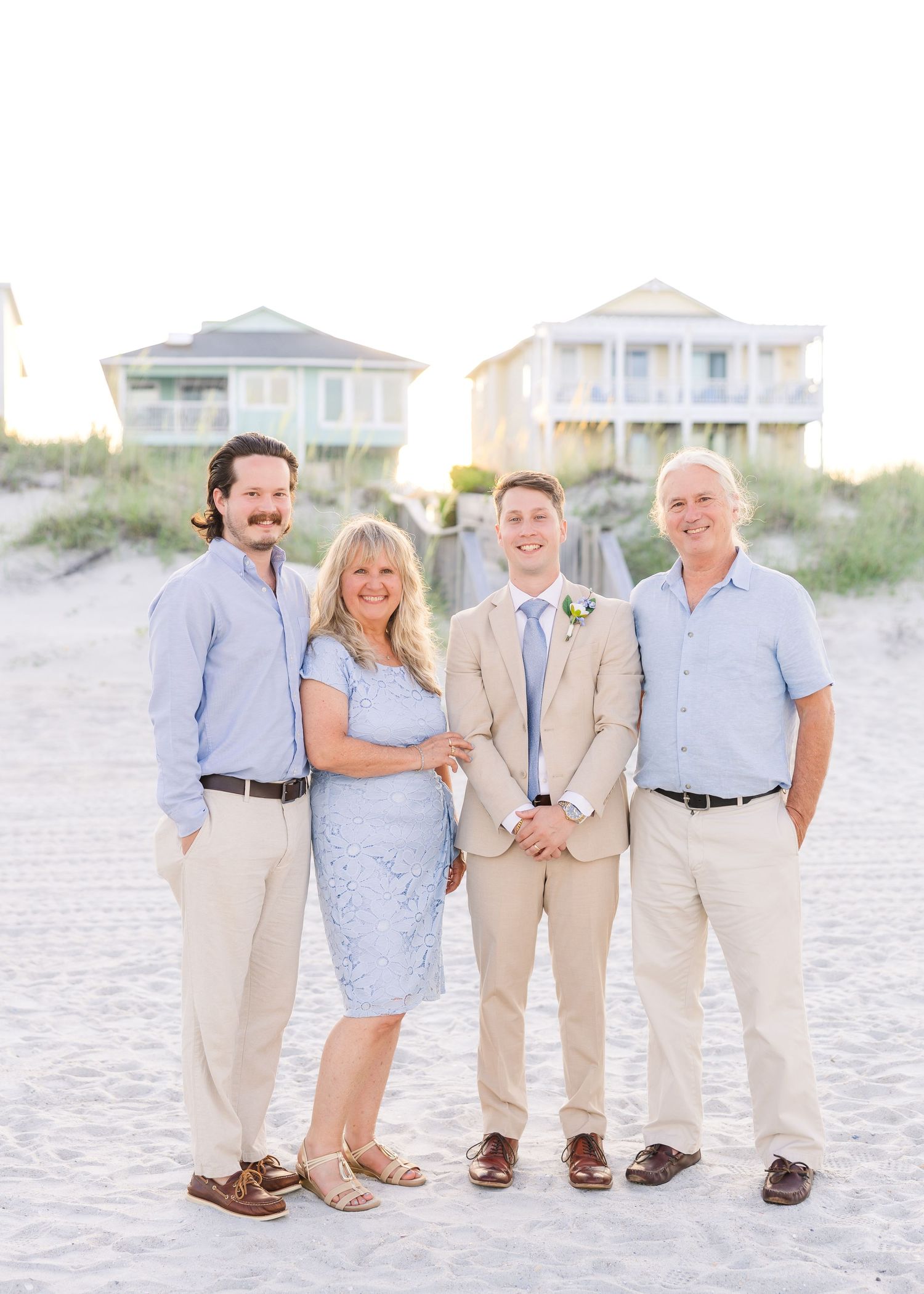 Groom poses with his family of four on sandy beach with colorful beach houses in the background in Carolina Beach near Wilmington, NC.