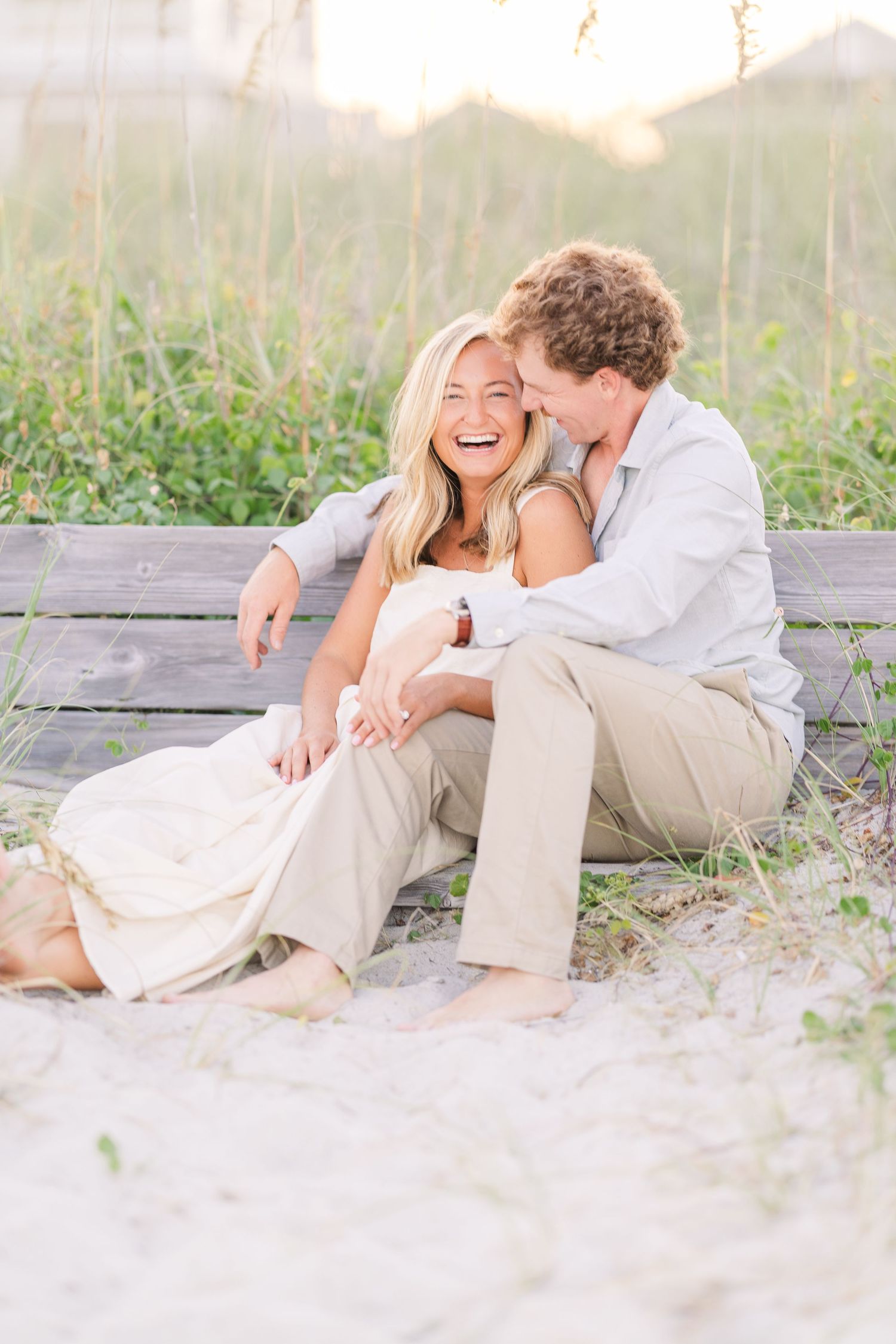 Couple laughing together while relaxing on wooden bench at beach during golden hour.