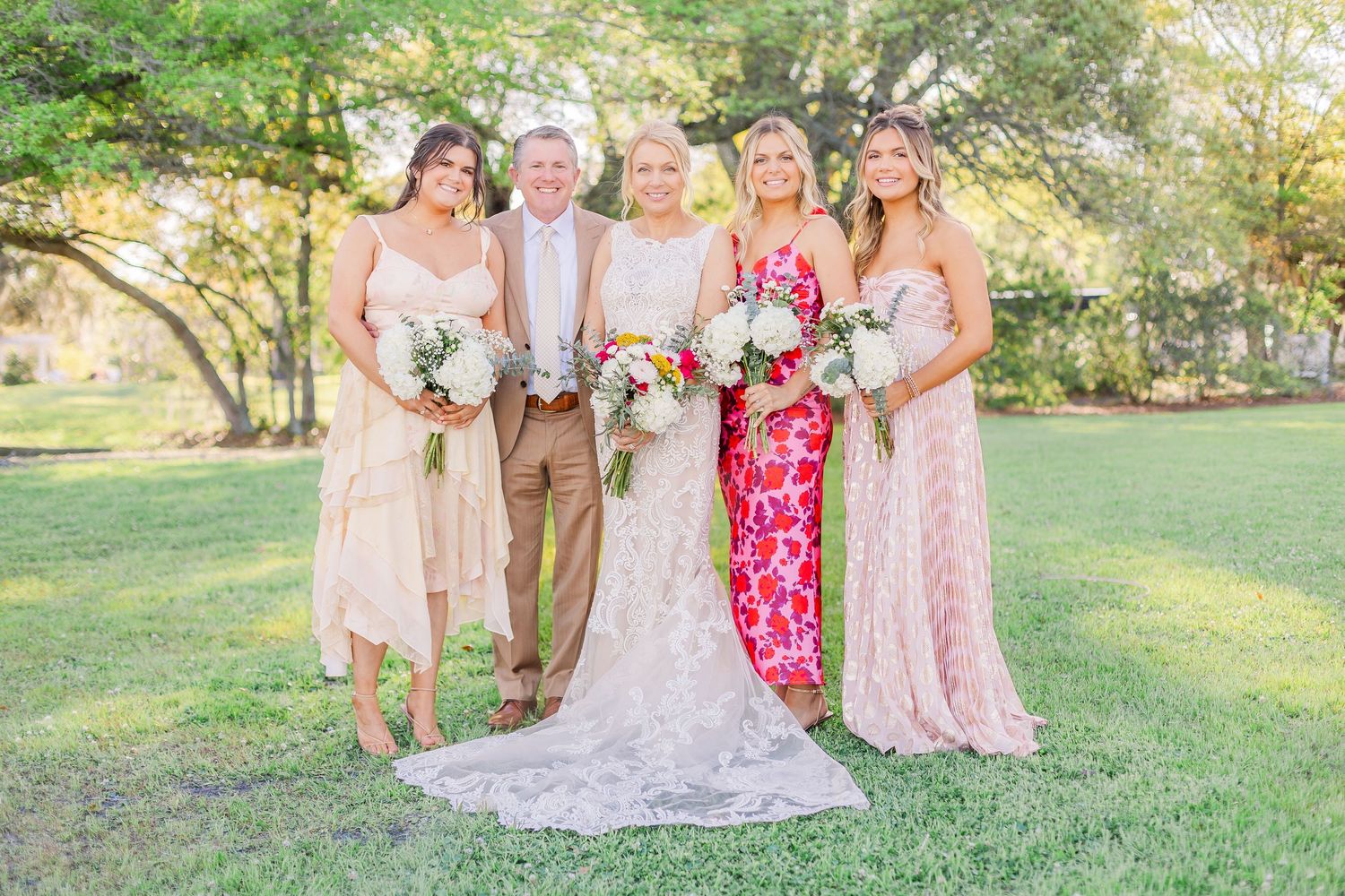 Wedding party gathers on grassy lawn with bridesmaids in pink and red dresses.