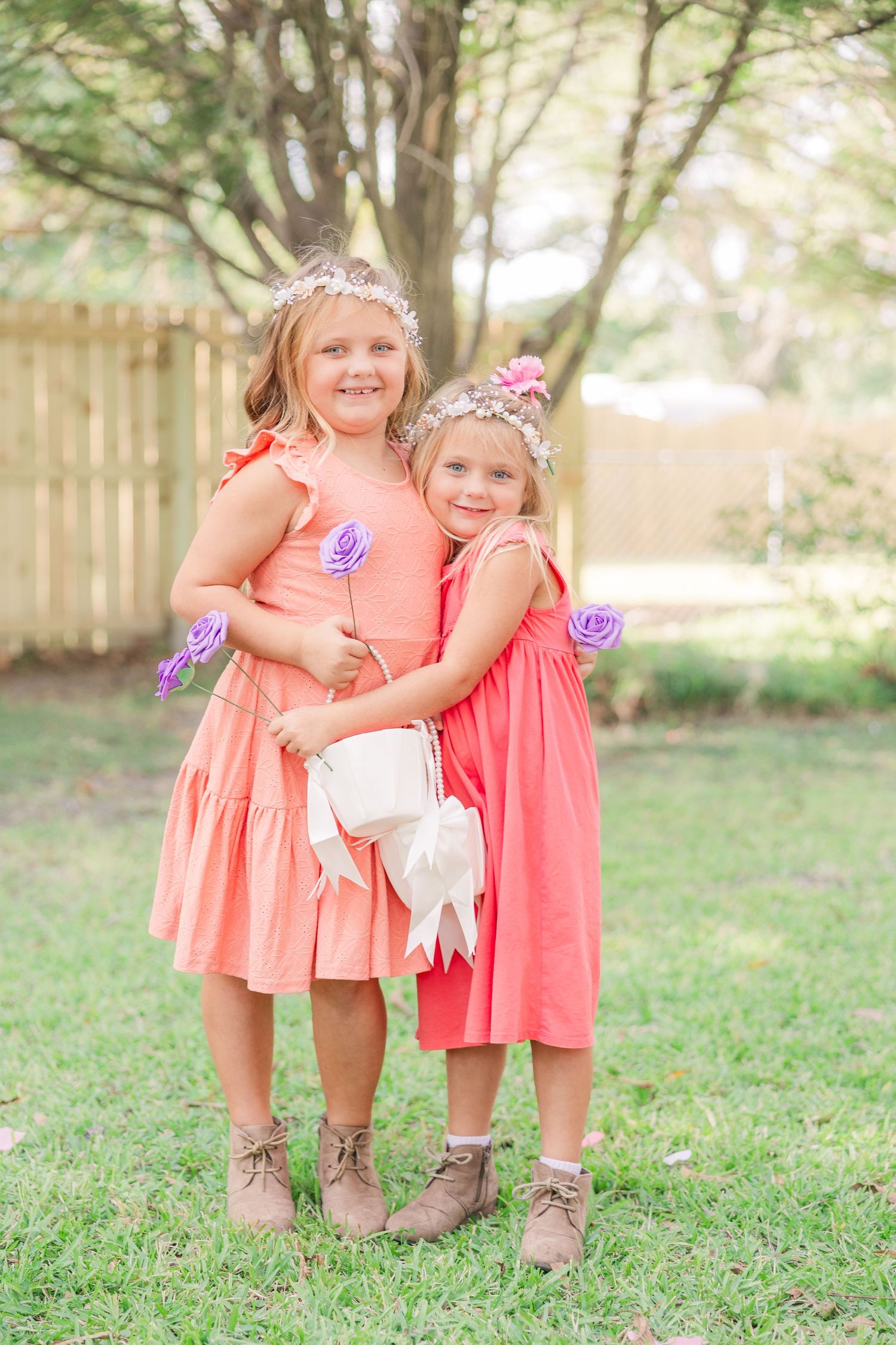 Young attendants in coral dresses and boots pose playfully on grassy lawn at outdoor wedding.