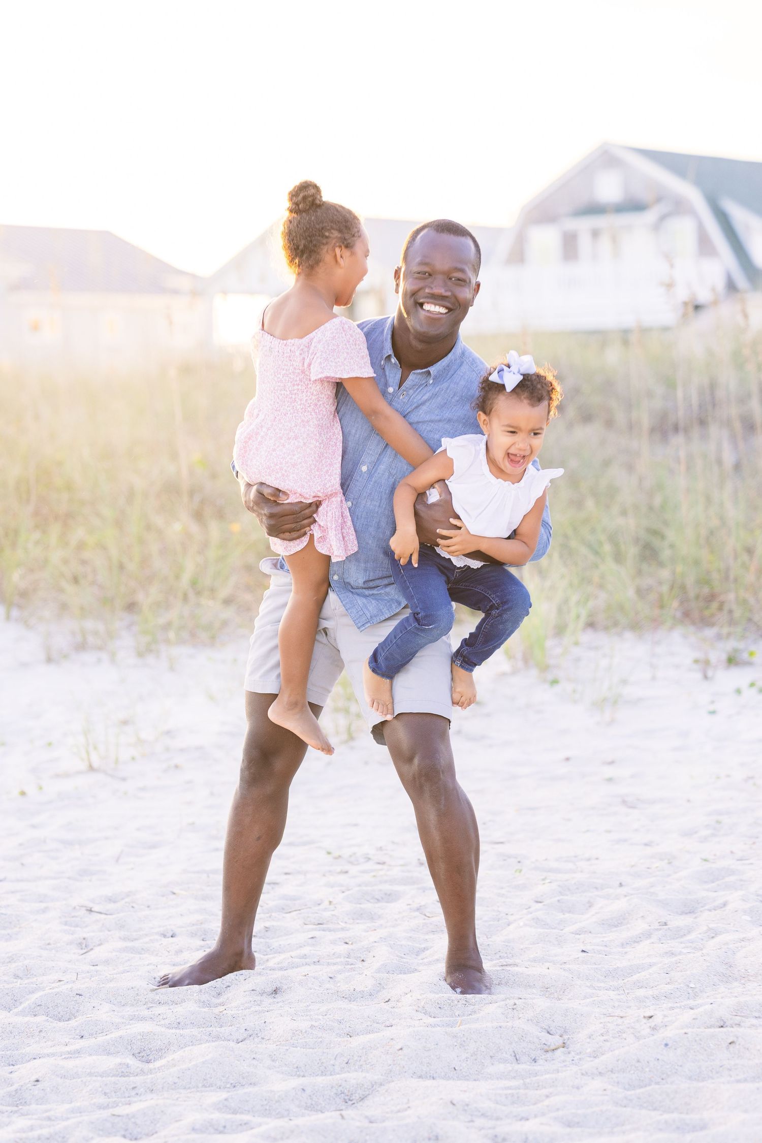 A joyful family moment on the beach as a parent playfully holds two children in casual summer attire.