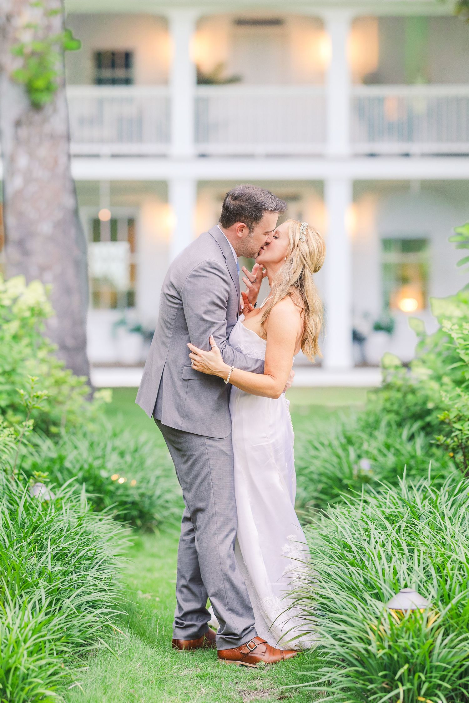 A romantic wedding couple shares a kiss in a garden setting with a white colonial style building in the background.