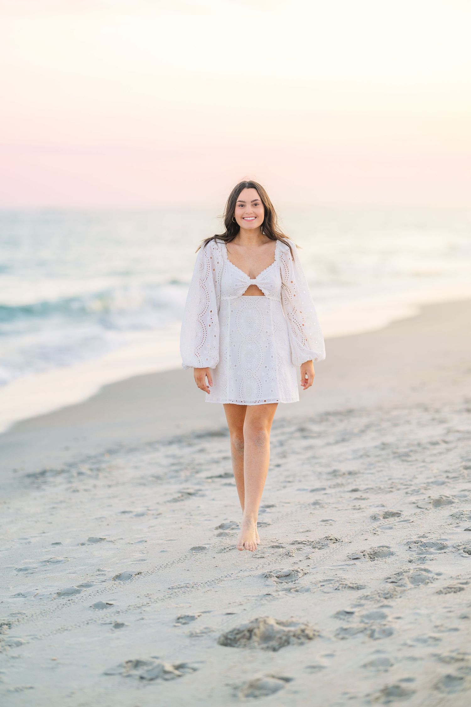 Barefoot beach walk in white cutout dress against pastel sunset sky.