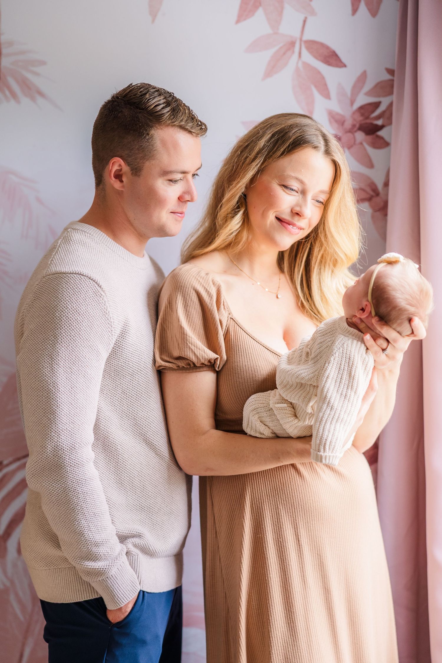 A tender family moment captured against a pink floral backdrop in soft natural light.