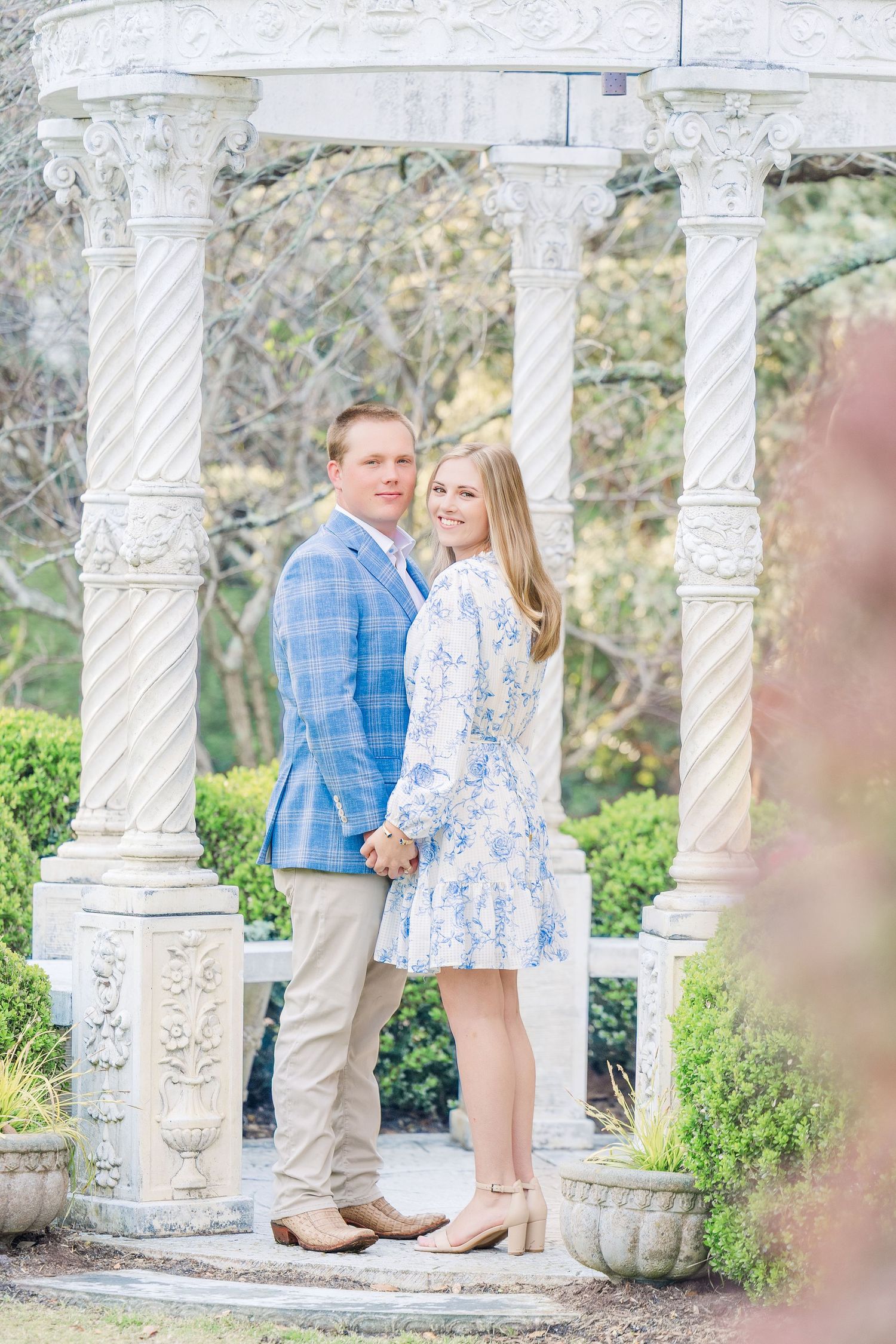 A couple stands together among ornate white stone columns in a garden setting during an engagement photoshoot.