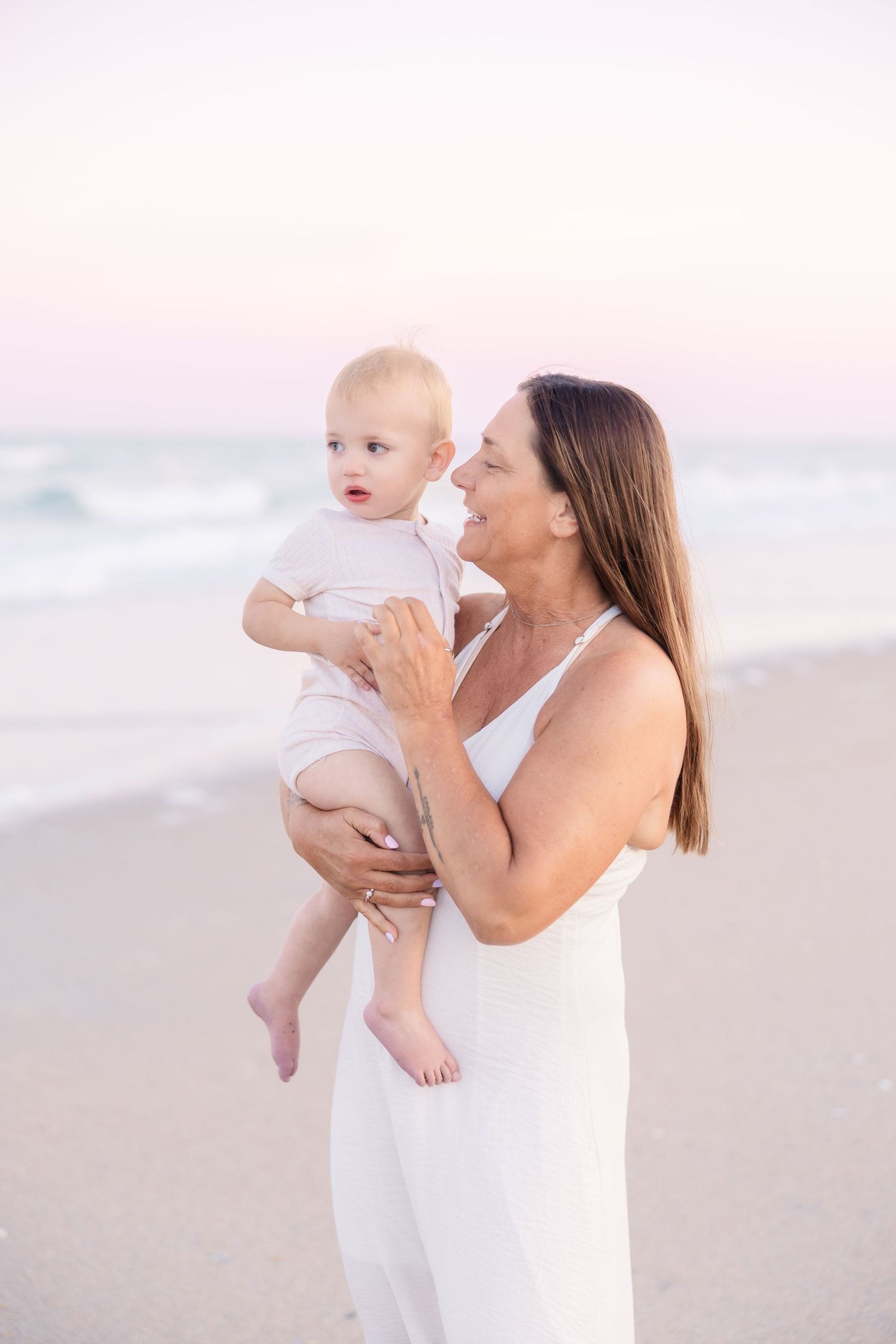A smiling woman in a white dress holds a happy baby on a beach at sunset.