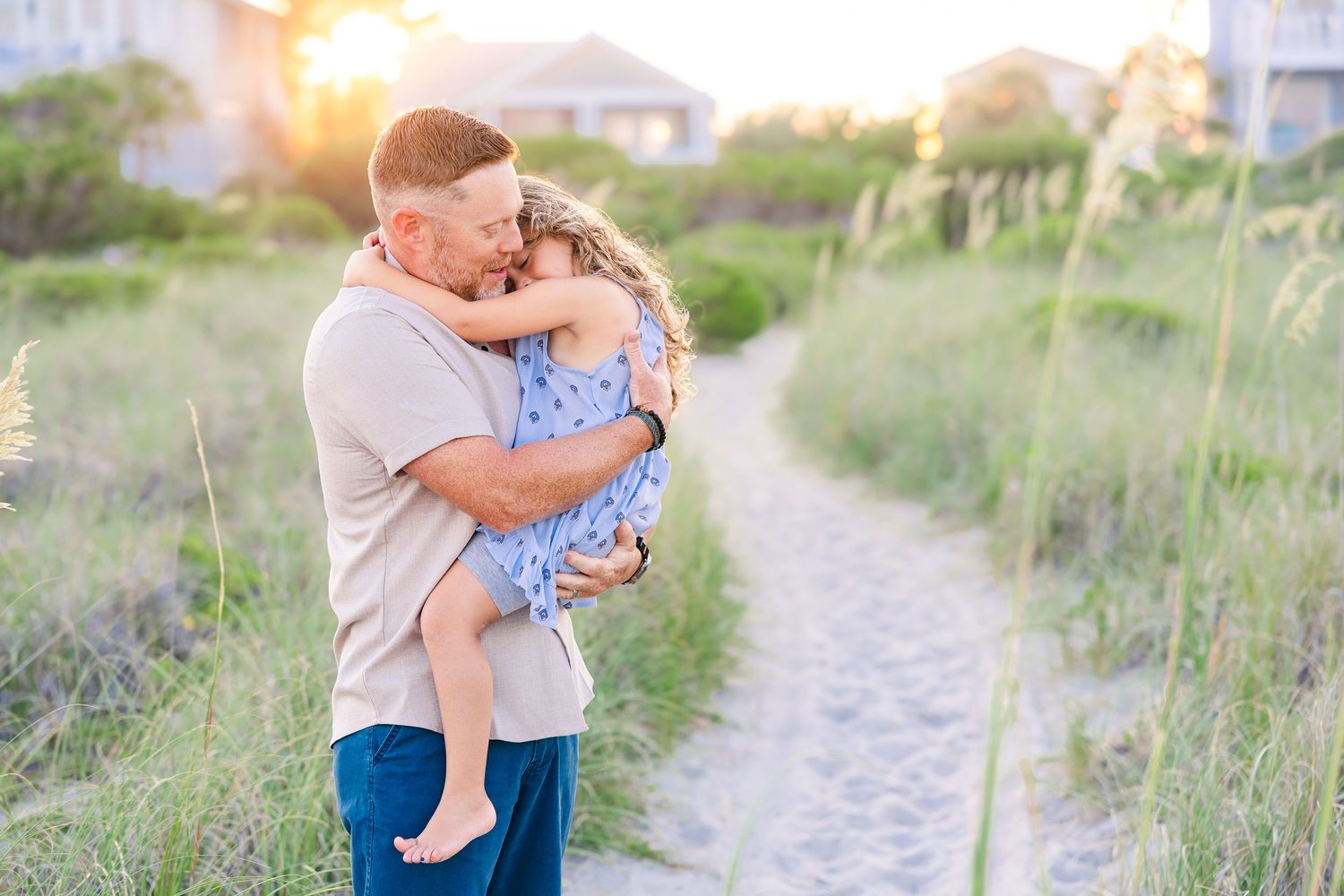 A father lovingly embraces his young daughter on a sunny beach path surrounded by sea oats at golden hour.