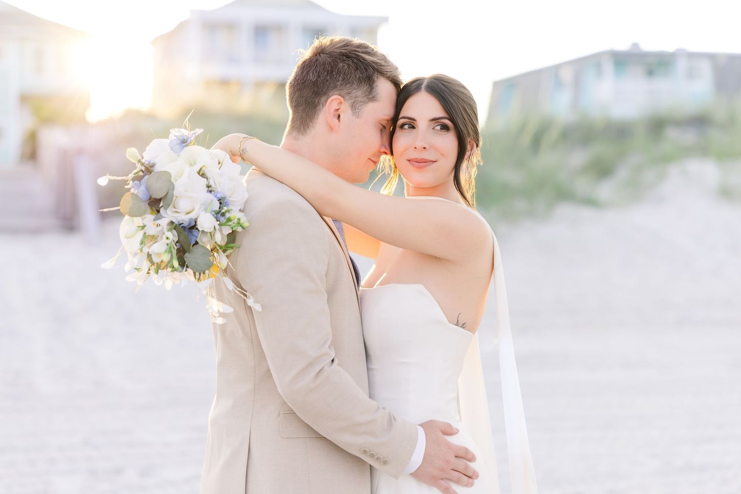 Bride and groom embrace on a sunlit beach, she holds white bouquet, wearing strapless gown, he in tan suit in Carolina Beach near Wilmington