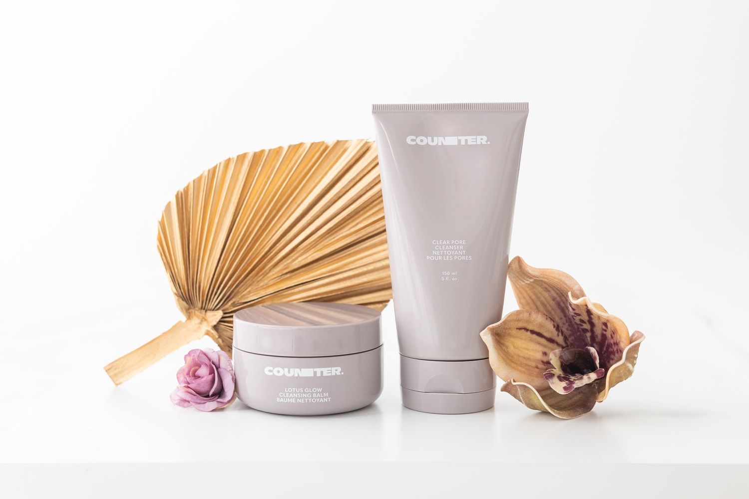 Minimalist beauty product flatlay with dried palm leaves and purple flowers against white background.