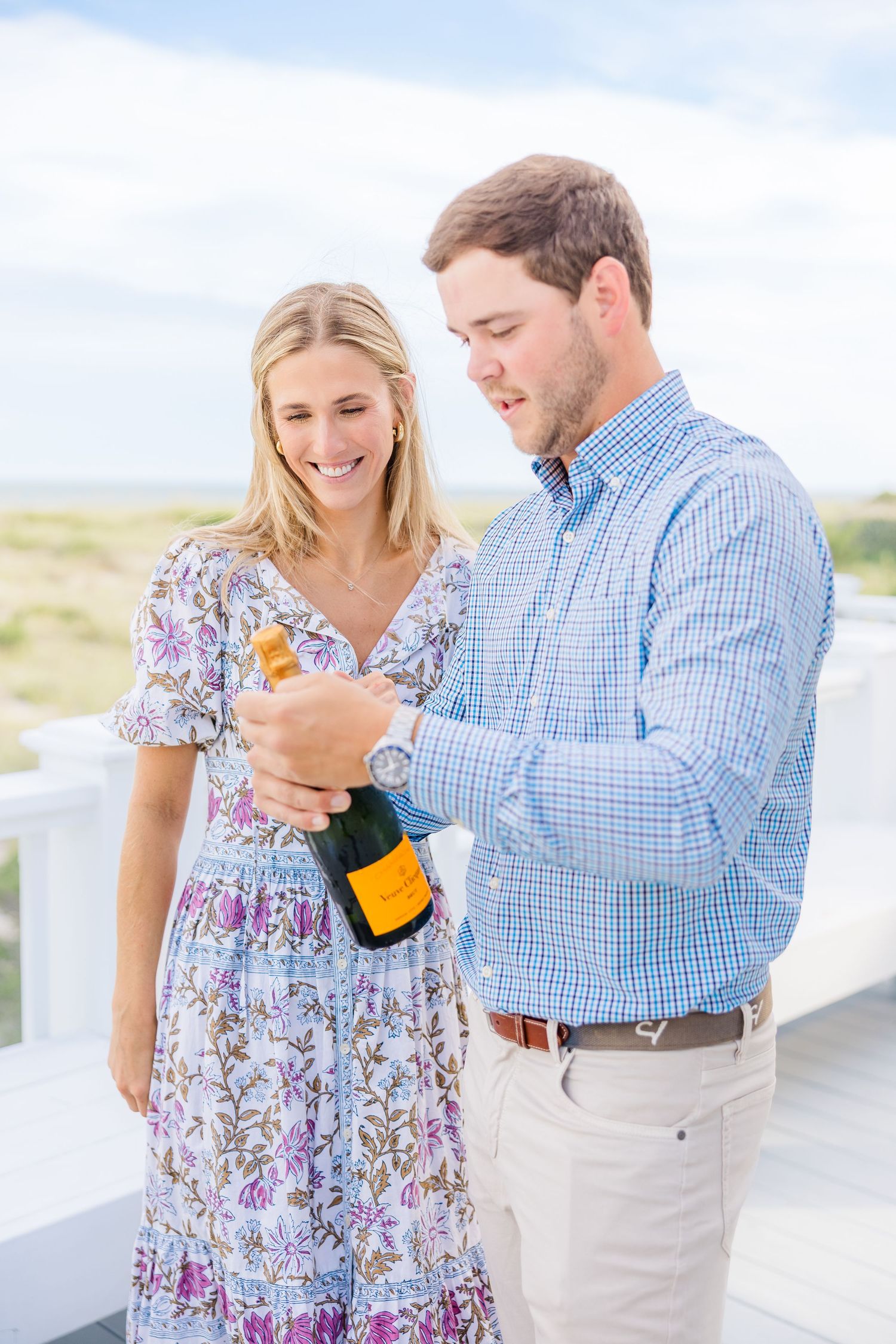 A couple celebrates with champagne on a coastal porch wearing blue and floral outfits.