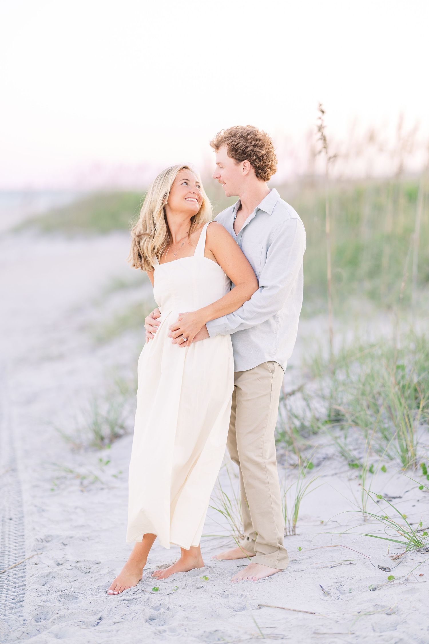 Playful couple in white and khaki outfits dancing on beach with sea grass in background.