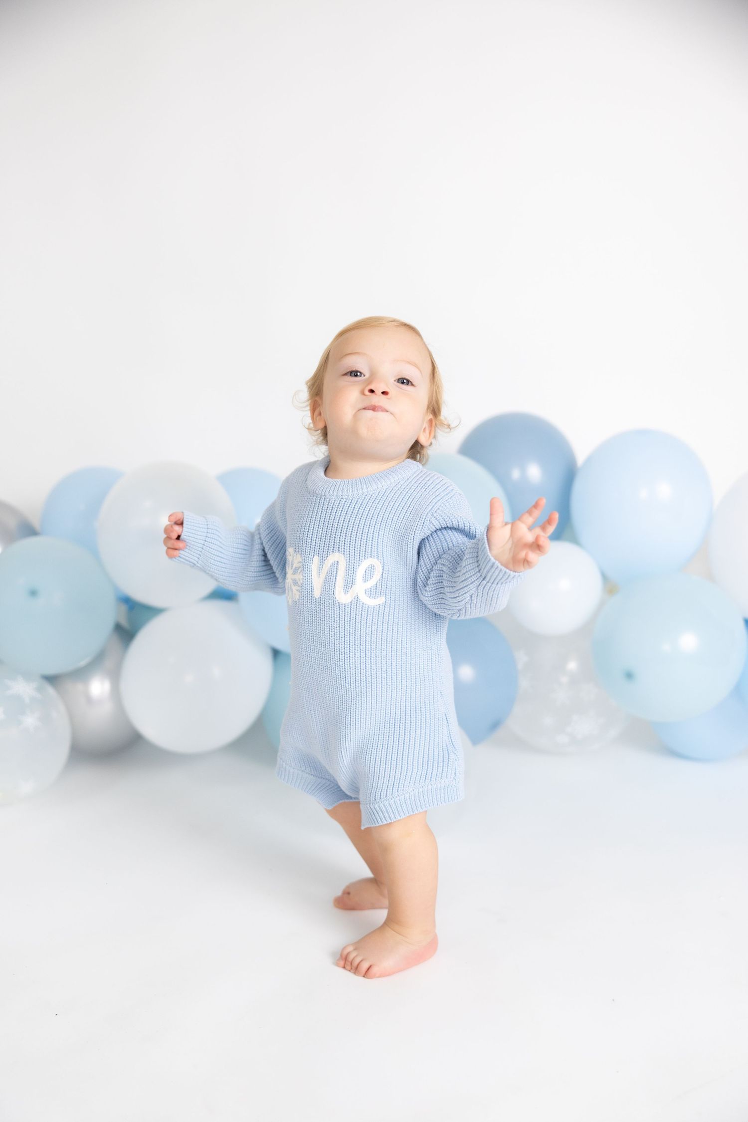 Baby in pale blue outfit stands against white backdrop with blue balloon garland.