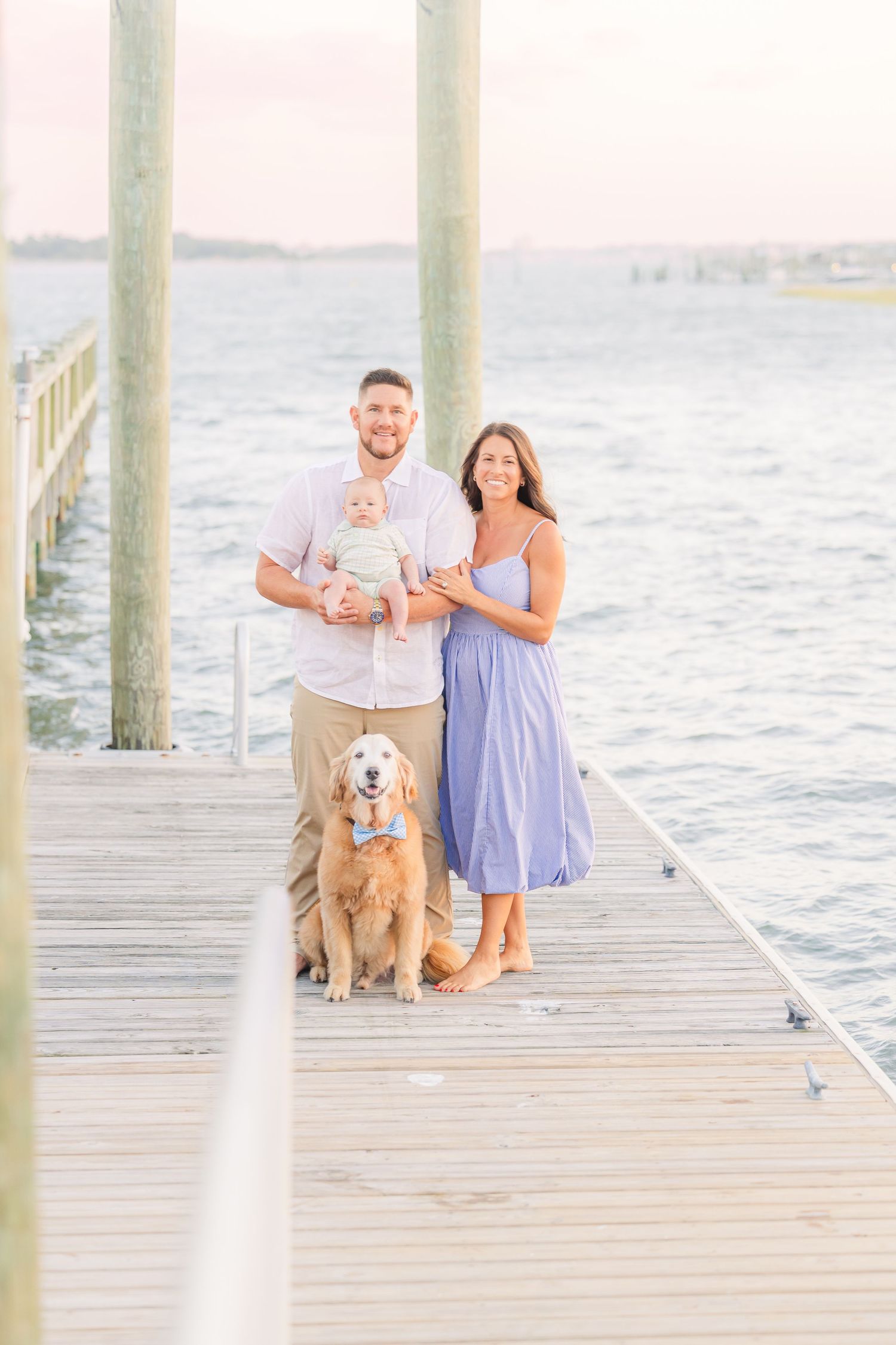 A family poses with their golden retriever on a waterfront pier during sunset.