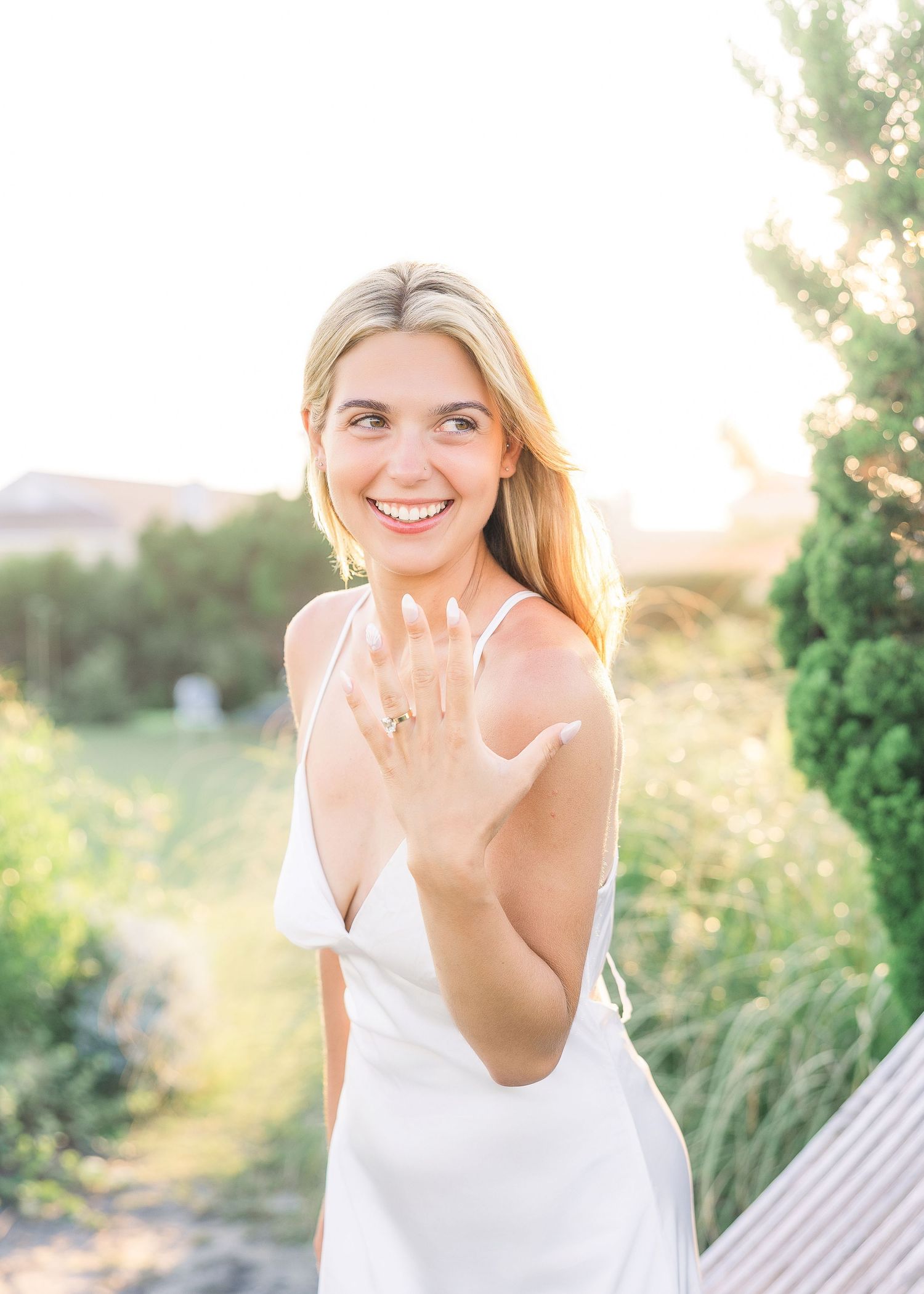 Smiling blonde woman in white dress showing engagement ring outdoors in golden hour light.