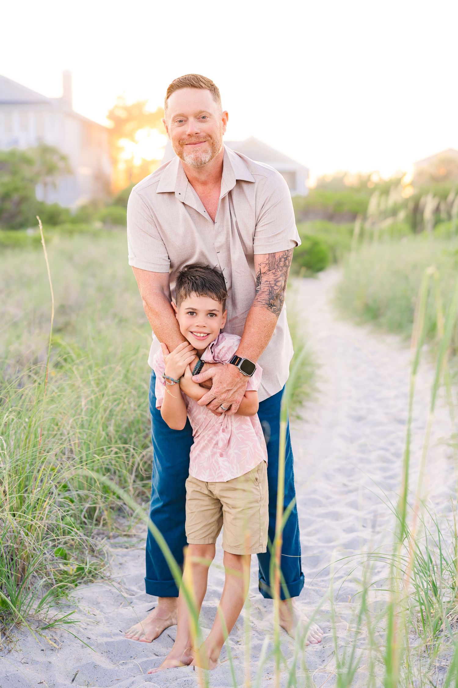 A father hugs his young son on a sandy beach path at sunset, both smiling with dune grass around them.