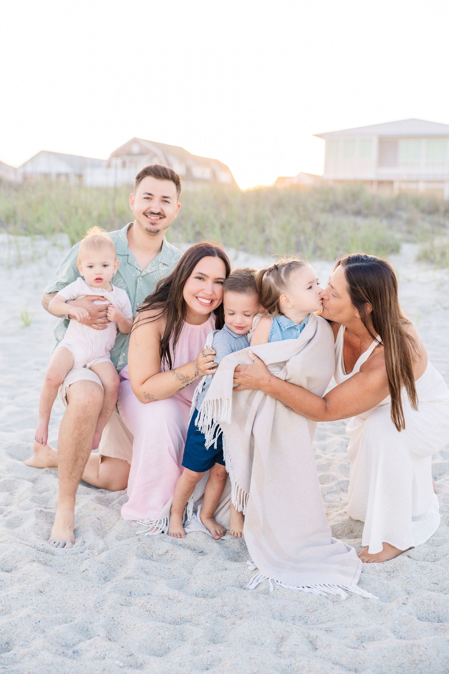 Happy extended family sits and cuddles together on sandy beach during warm sunset photography session.