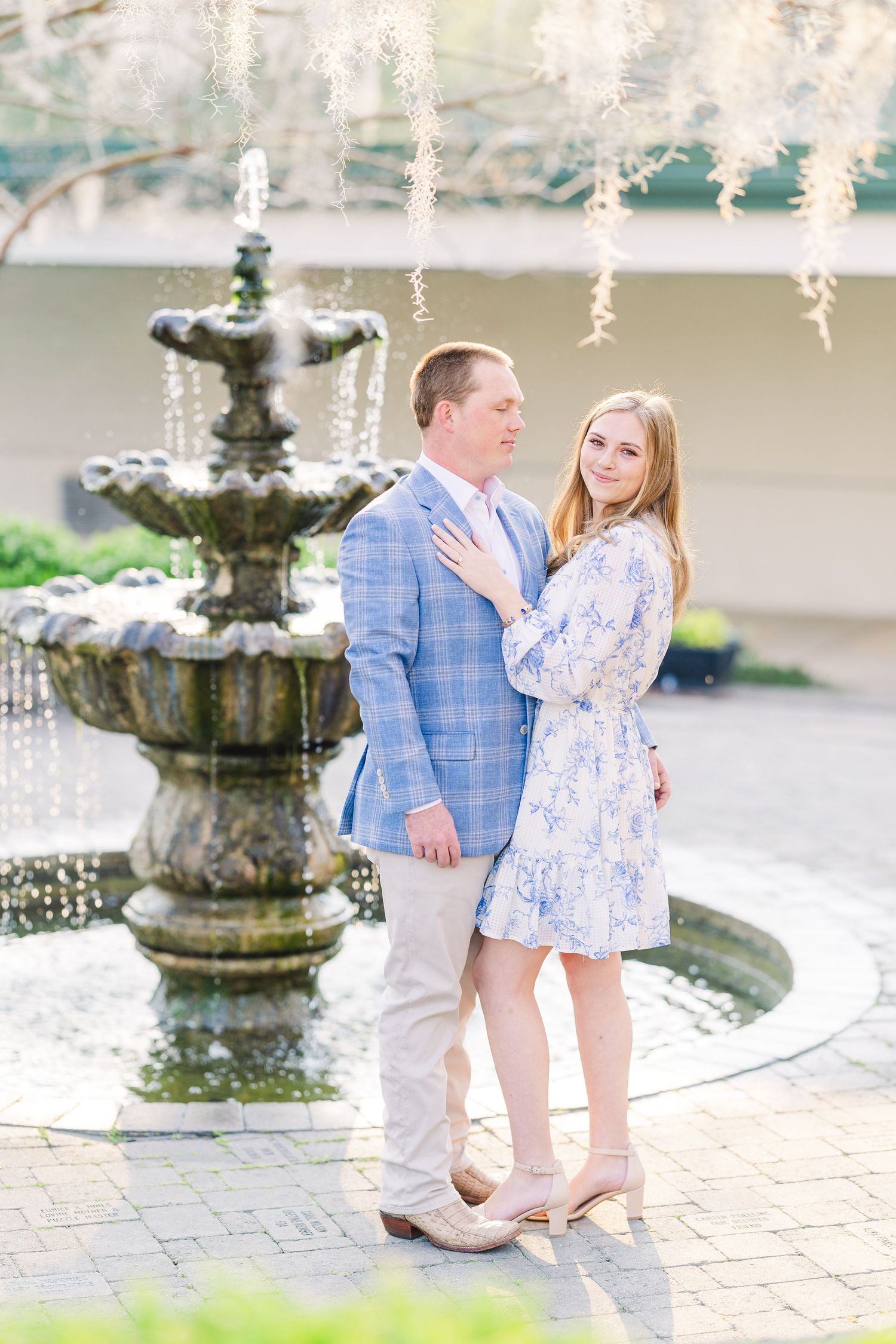 A couple poses together by an ornate stone fountain in a garden setting with hanging string lights above.