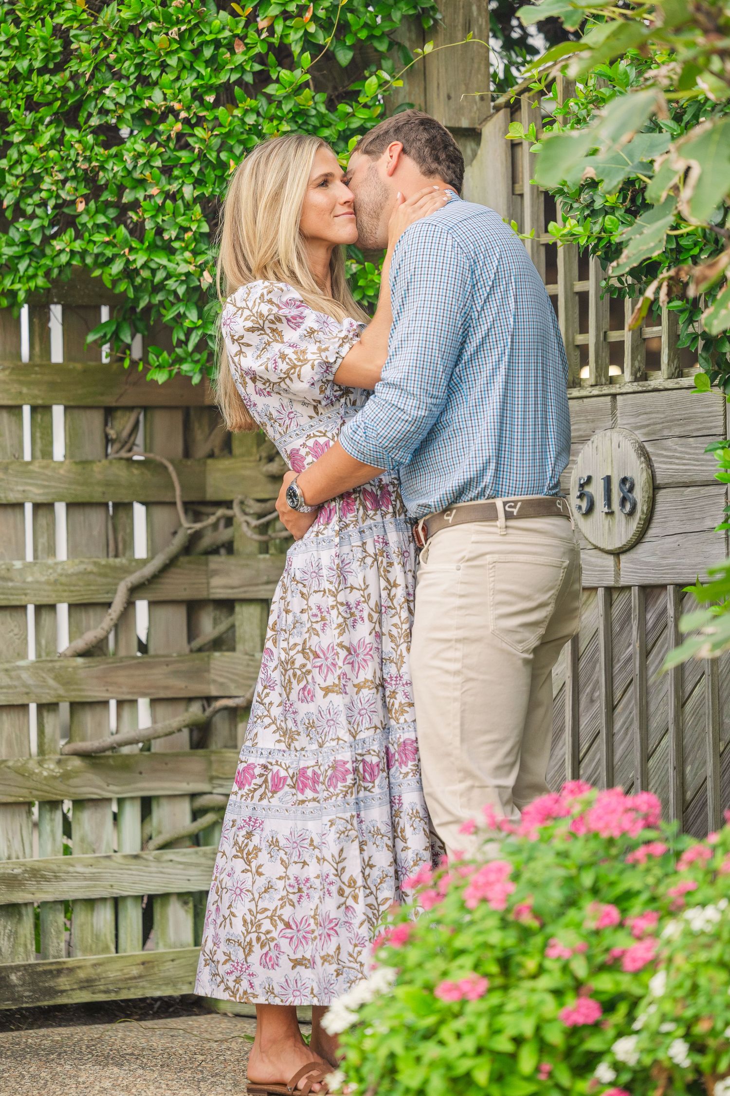 A couple embraces by a garden fence with blooming pink flowers and greenery.