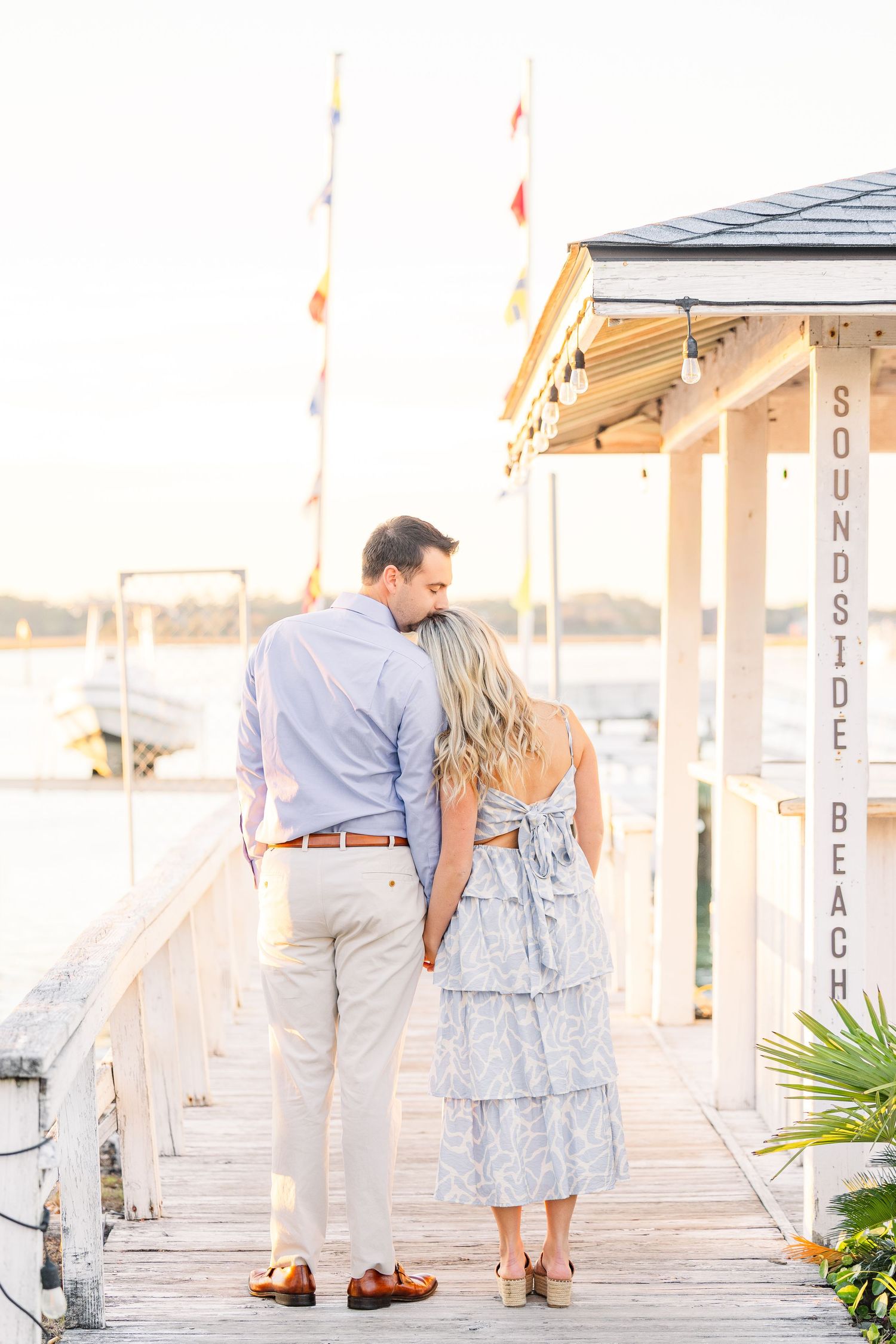 Couple holding hands while standing on wooden pier with coastal architecture in background.