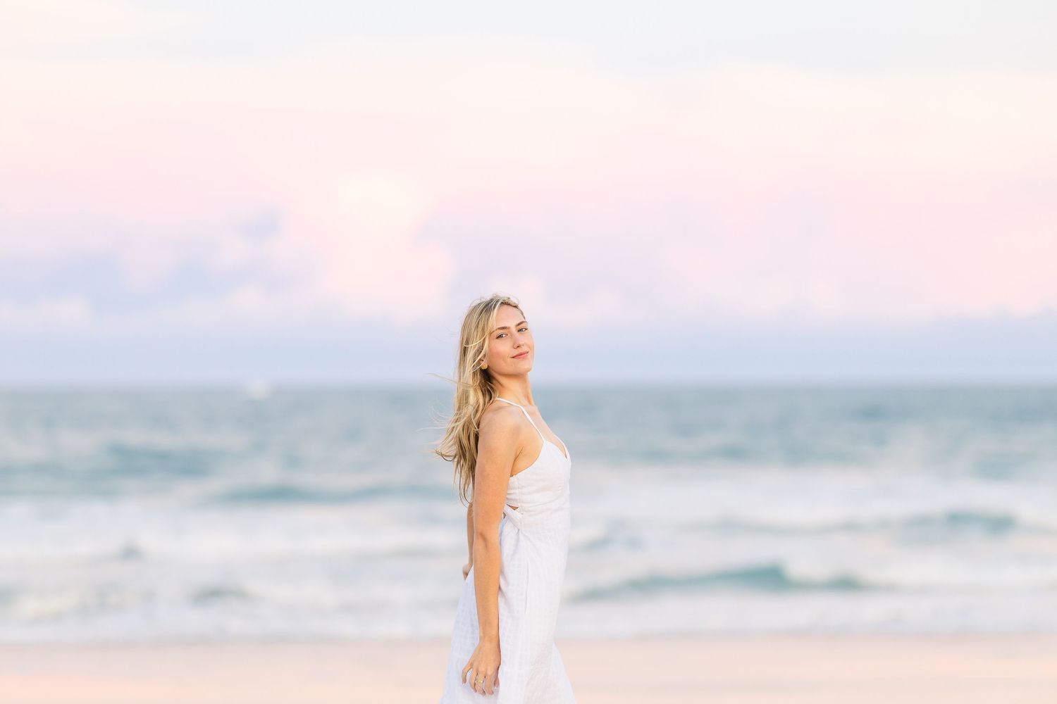 A person in a flowing white dress stands on a sandy beach during golden hour with pastel skies.