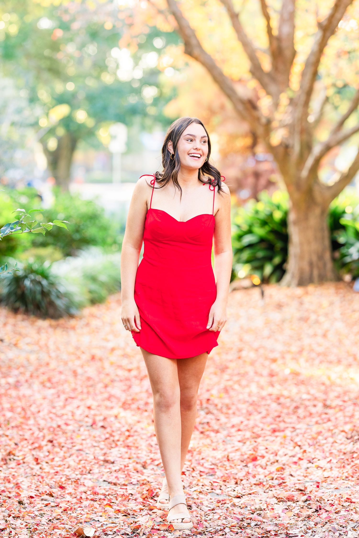 Playful autumn photoshoot in a red dress among fallen leaves and trees.