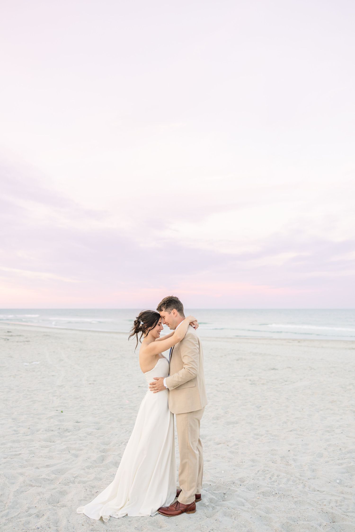 Romantic couple sharing an intimate moment on a beach at dusk, bride in flowing white dress and groom in beige suit in Carolina Beach