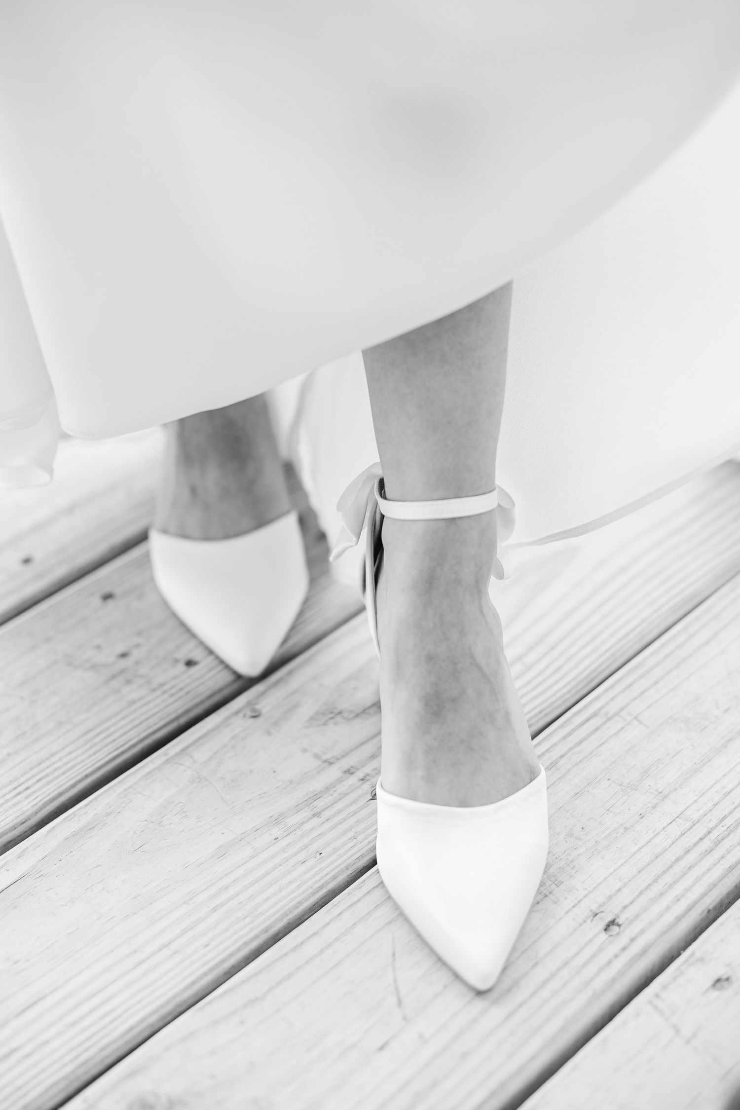 Black and white close-up of bride wearing classic white pointed-toe heels with ankle straps on wood in Carolina Beach near Wilmington, NC.