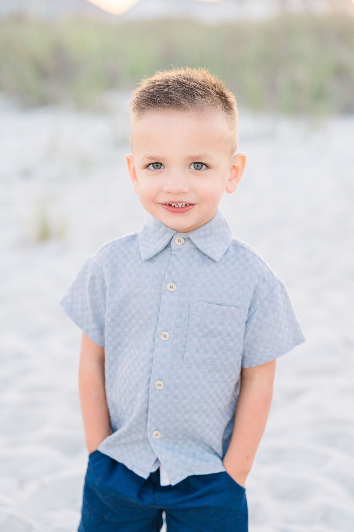 Smiling young boy in a light blue button-up shirt standing on a sandy beach.