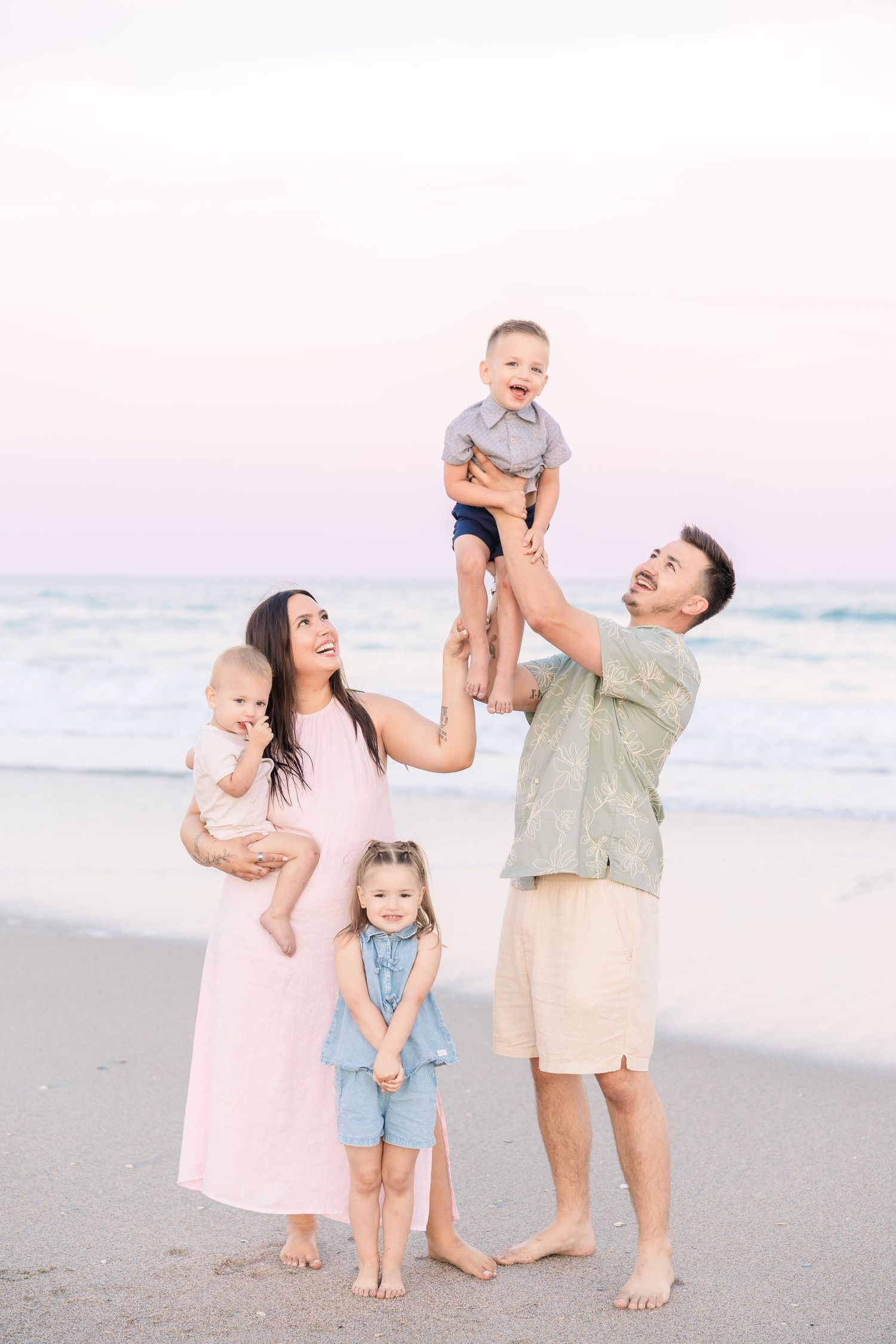 Family of five enjoying a beach sunset, dad lifting toddler overhead while mom holds baby and daughter stands smiling.