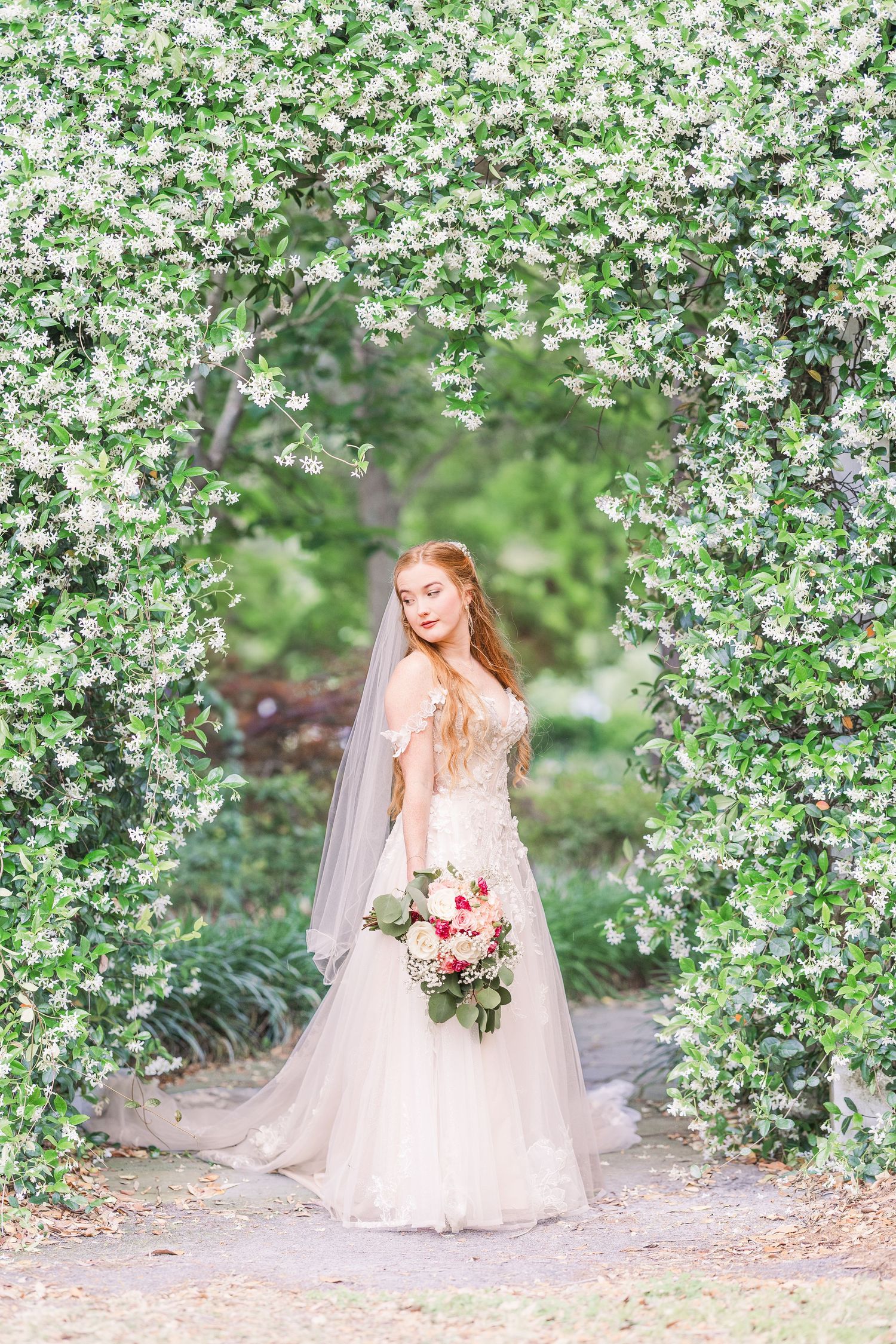 Bride in flowing white gown poses beneath blooming white cherry blossoms holding a pink bouquet in harborway gardens in Wrightsville Beach,