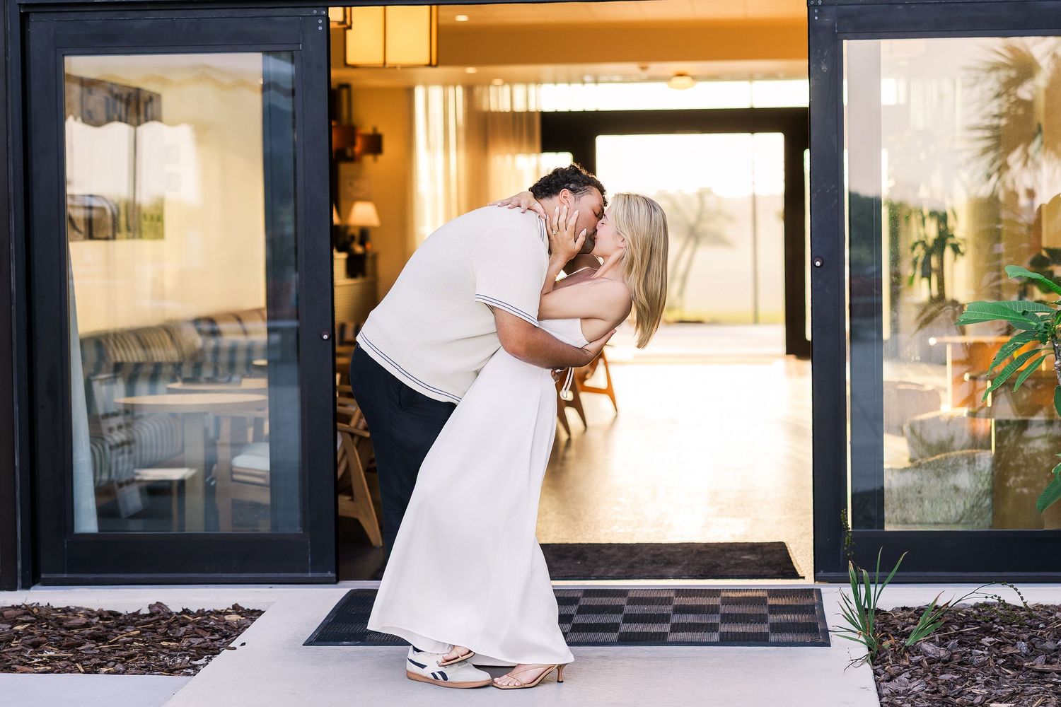 Couple sharing a romantic dip kiss in a doorway, woman in white dress and heels at golden hour.