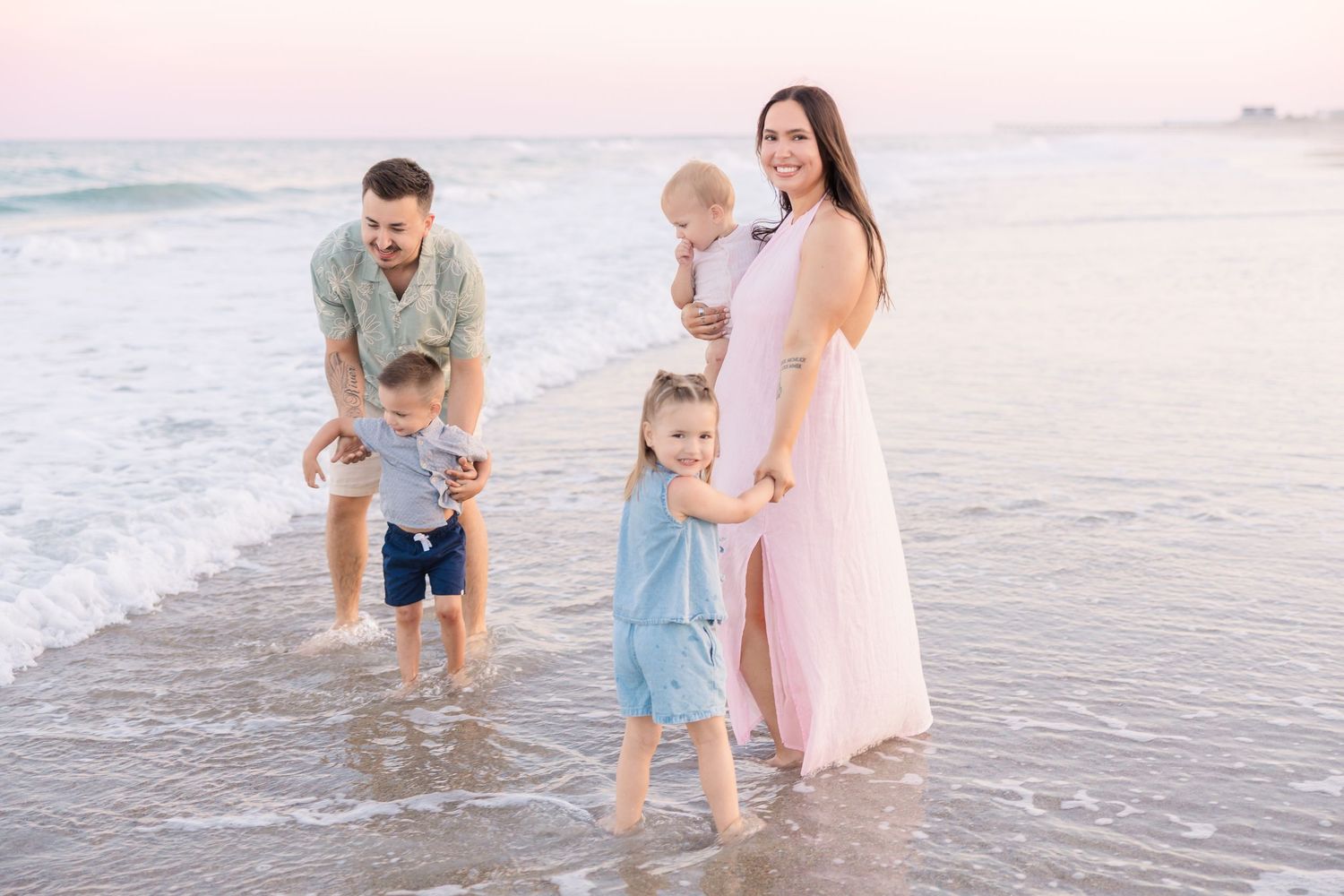 A joyful family of five plays together in the ocean waves at a colorful beach sunset.