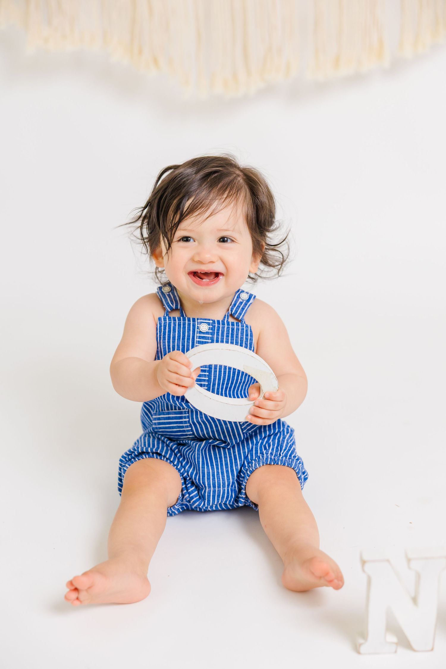Smiling baby in blue striped romper sits on white floor holding letter C during first birthday photo session.